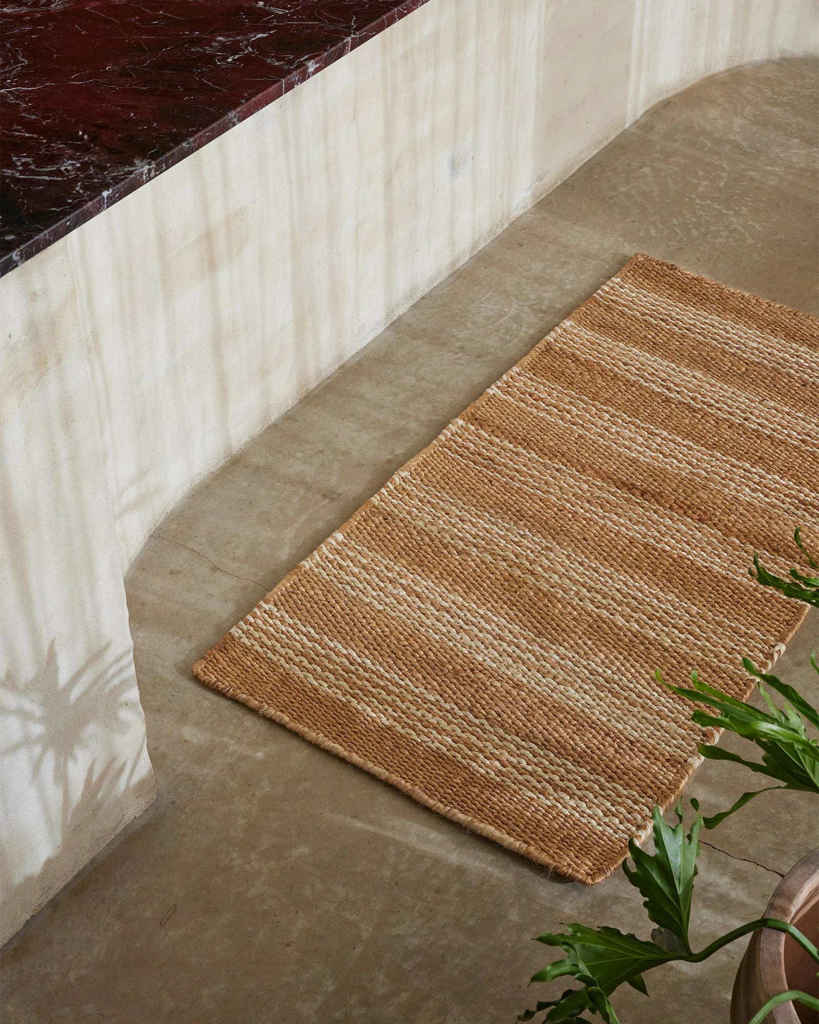 Natural jute door mat layered in neutral minimalist entryway with cream textiles and deep burgundy marble, styled with fresh green foliage