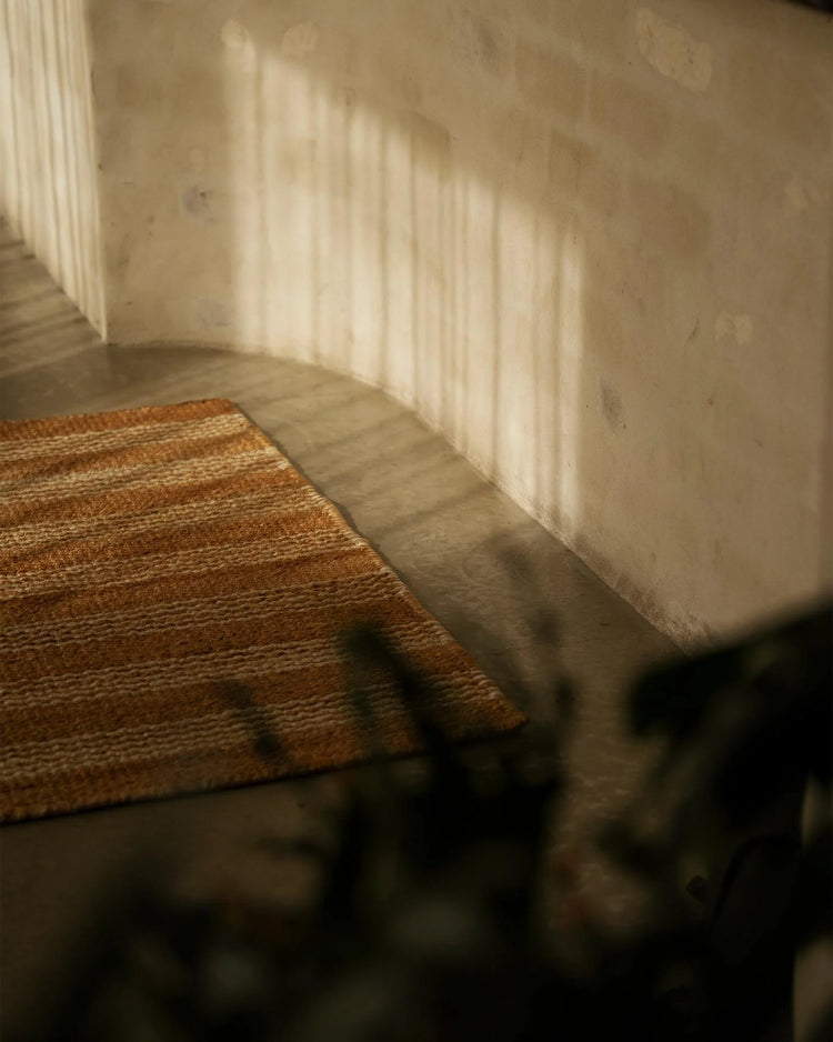 Natural jute doormat in warm terracotta tones placed at entryway, bathed in soft window light casting linear shadows on cream walls, minimalist aesthetic