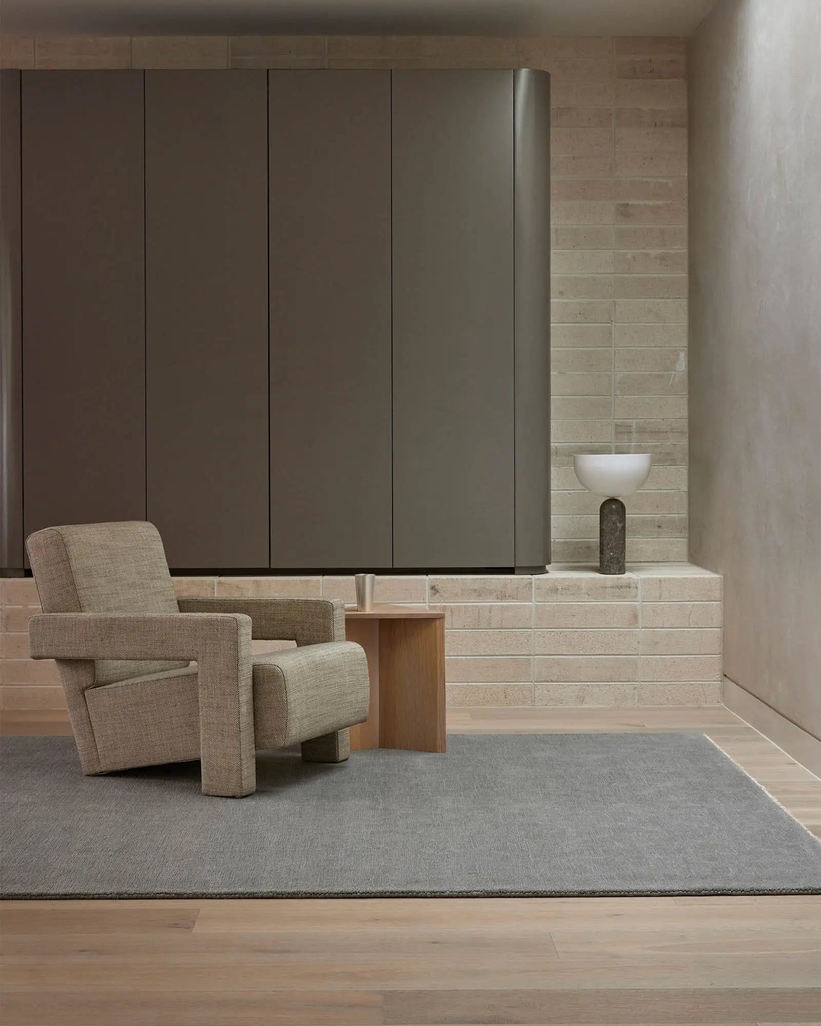 Neutral minimalist interior with warm beige linen chair on soft grey wool rug, taupe brick walls, warm wood accents and cool-toned cabinetry