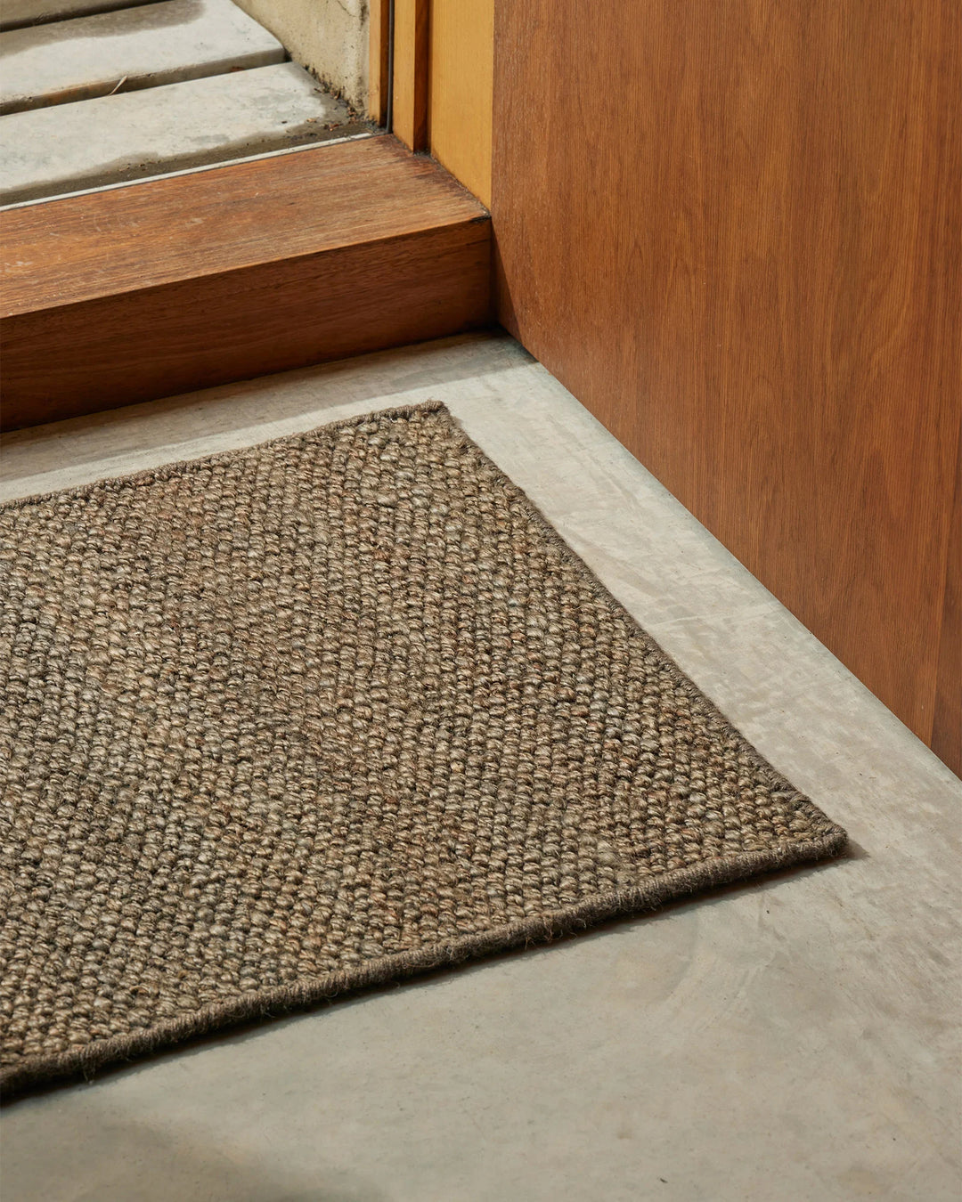 Natural jute doormat in earthy taupe placed at timber-framed entrance with pale stone flooring and warm wood panelling, creating textured, neutral entryway styling