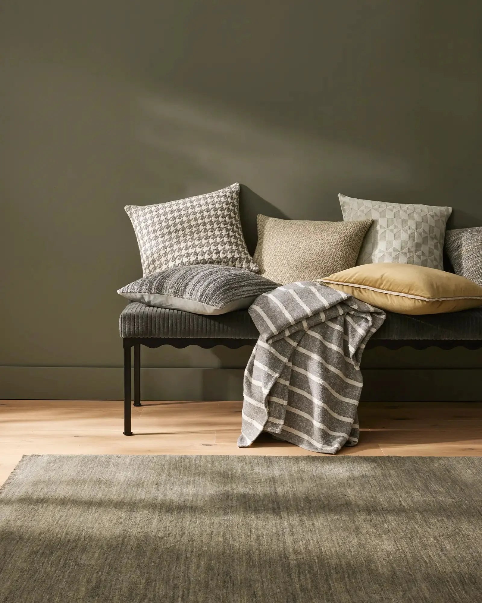 Sage green bedroom with layered textured pillows and striped linen throw on modern bench, neutral palette of cream, taupe and grey against warm timber flooring