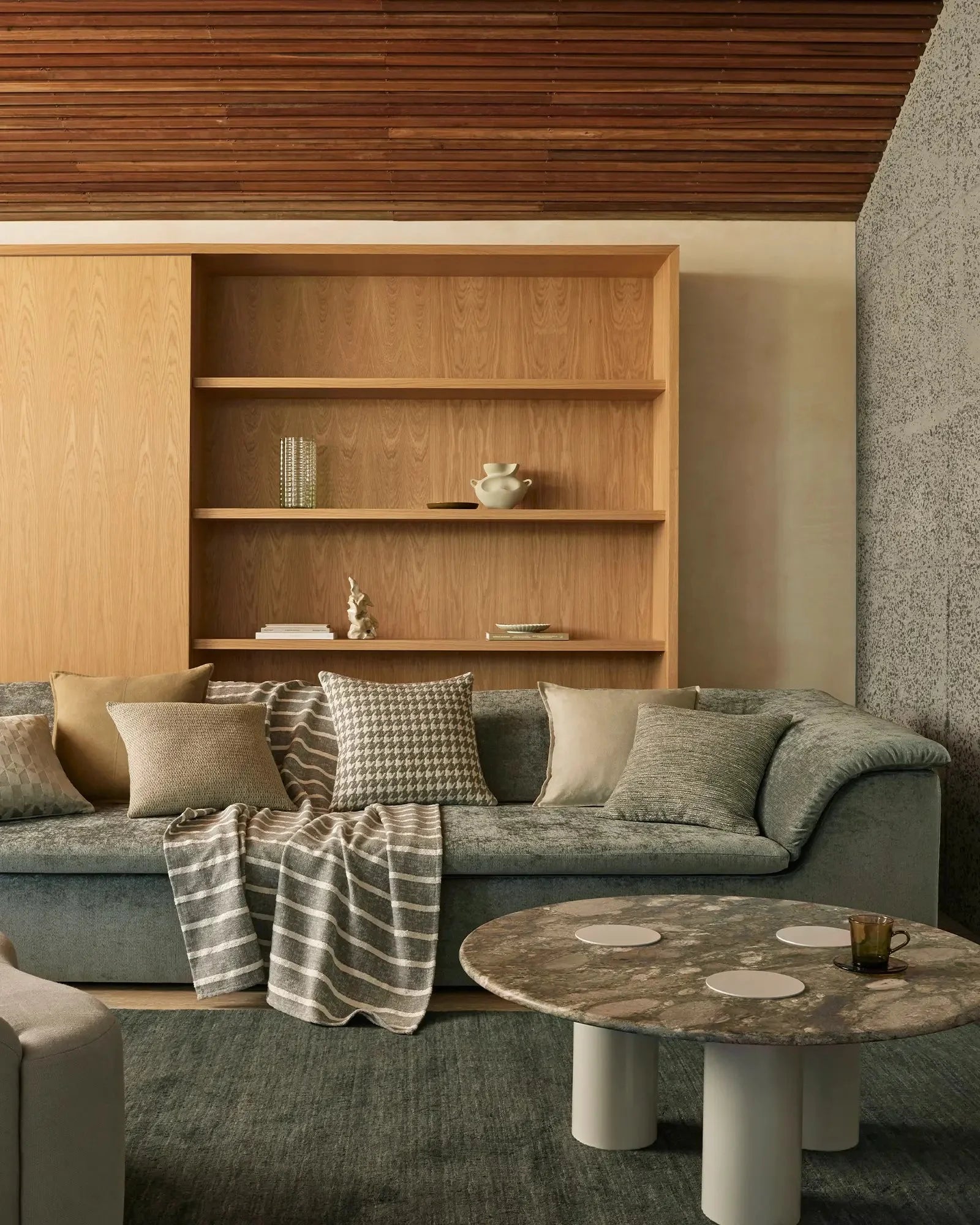 Warm minimalist living room with grey upholstered sofa, neutral linen and striped throws, geometric cushions in cream and sage, timber shelving, and marble-topped coffee table
