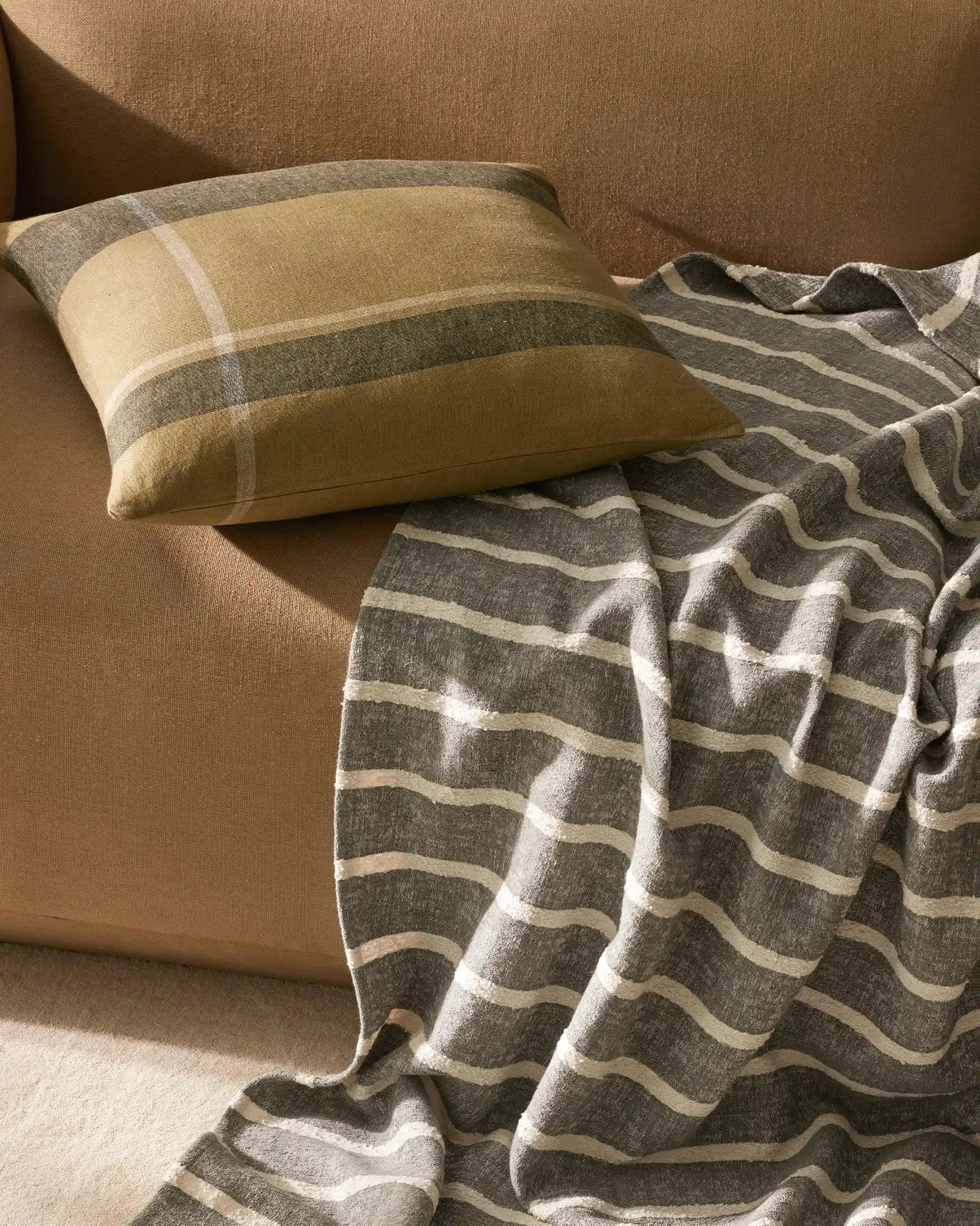 Warm neutral living room with earth-toned upholstery, layered striped throw in charcoal and cream, and coordinating cushion with horizontal colour blocking in sage and gold tones