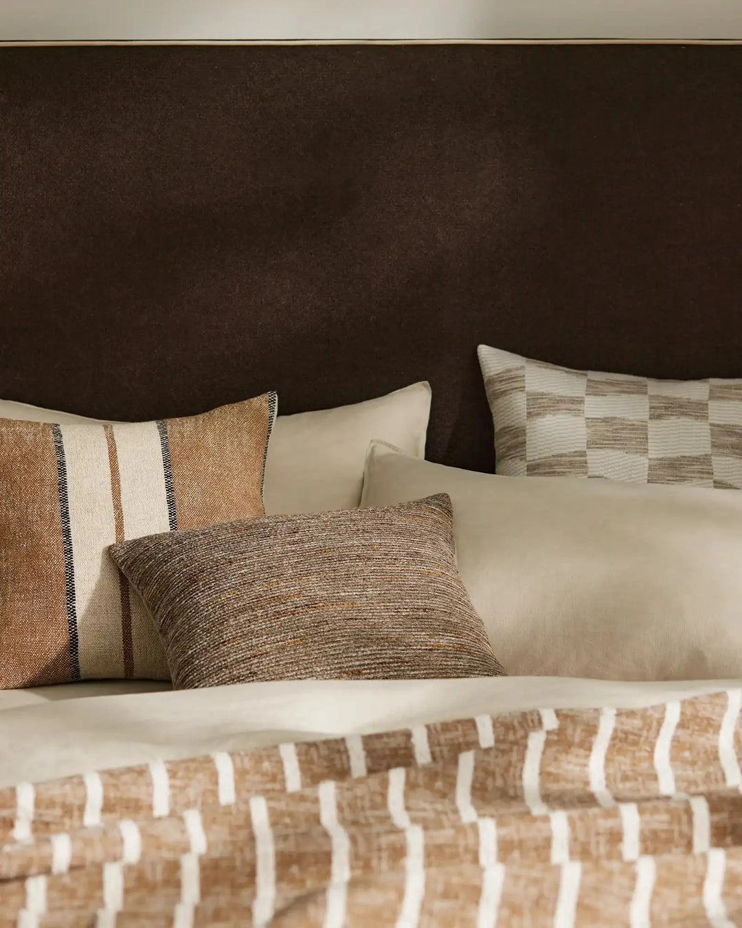Warm neutral bedroom styled with Vista Natural cushion and Ottavio Cocoa pillow, layered with textured throws in cream, tan and brown tones against chocolate headboard