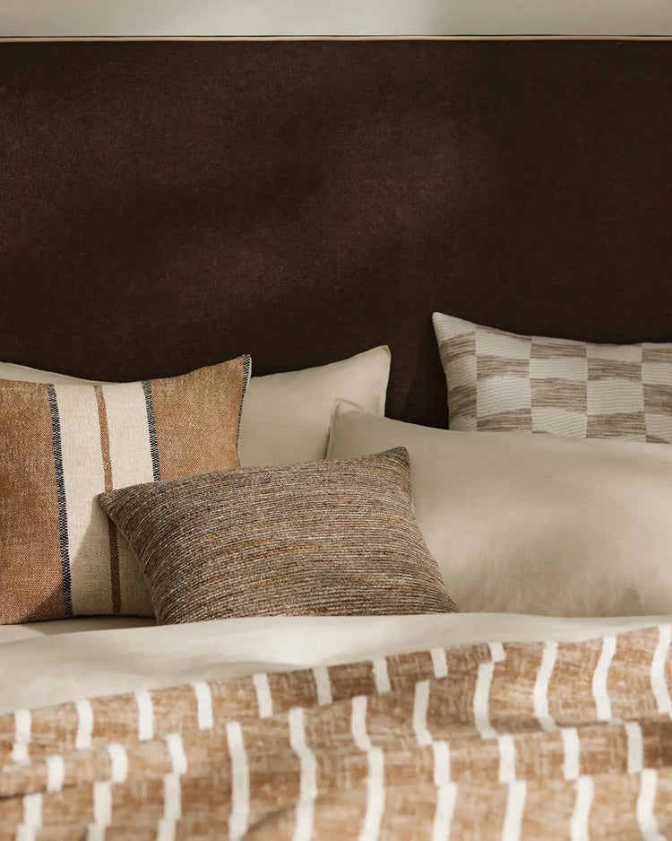 Warm neutral bedroom styled with Vista Natural cushion and Ottavio Cocoa pillow, layered with textured throws in cream, tan and brown tones against chocolate headboard