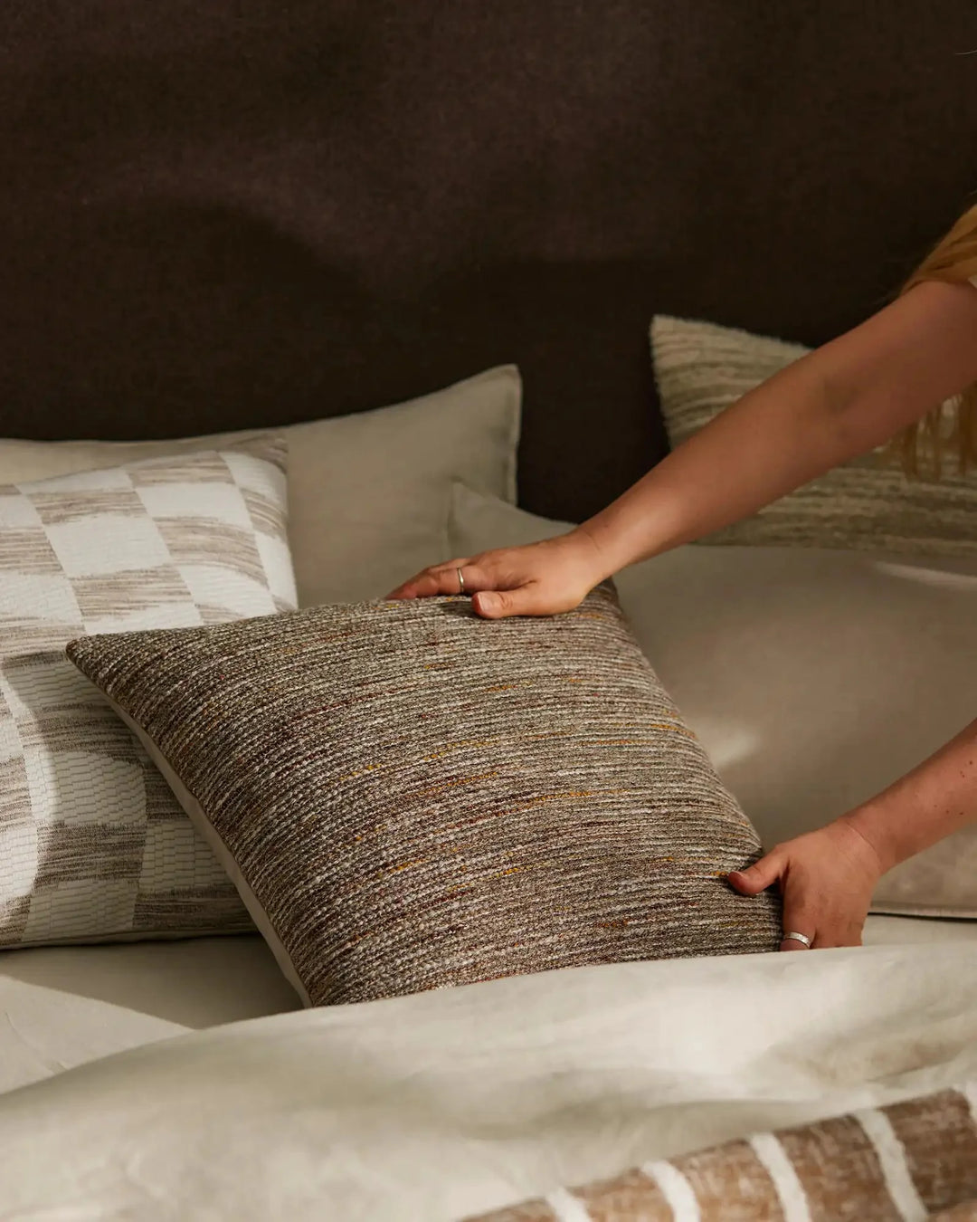 Hand arranging neutral linen and textured cushions on bed with warm taupe bedding, showcasing layered minimalist bedroom styling
