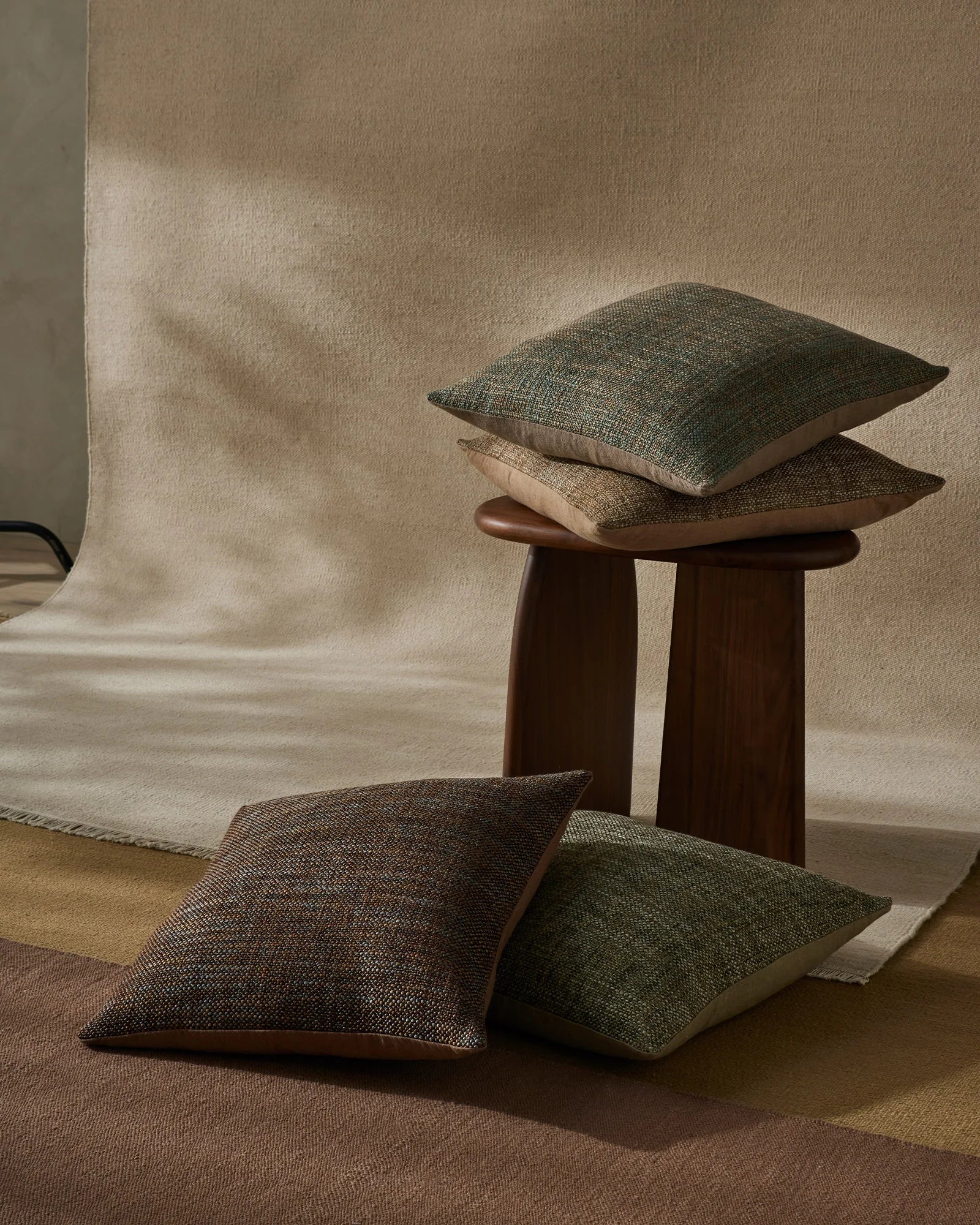 Earthy minimalist interior styling with stacked Castel cushions in moss, toffee and neutral tones, displayed on dark wood side table against soft linen backdrop