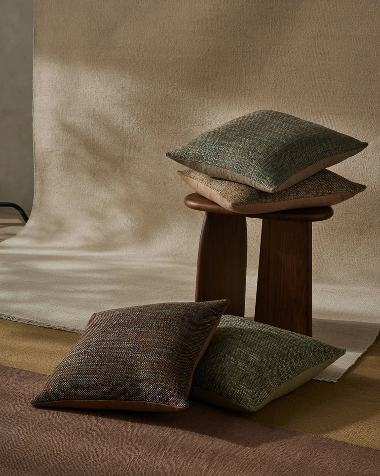 Earthy minimalist interior styling with stacked Castel cushions in moss, toffee and neutral tones, displayed on dark wood side table against soft linen backdrop