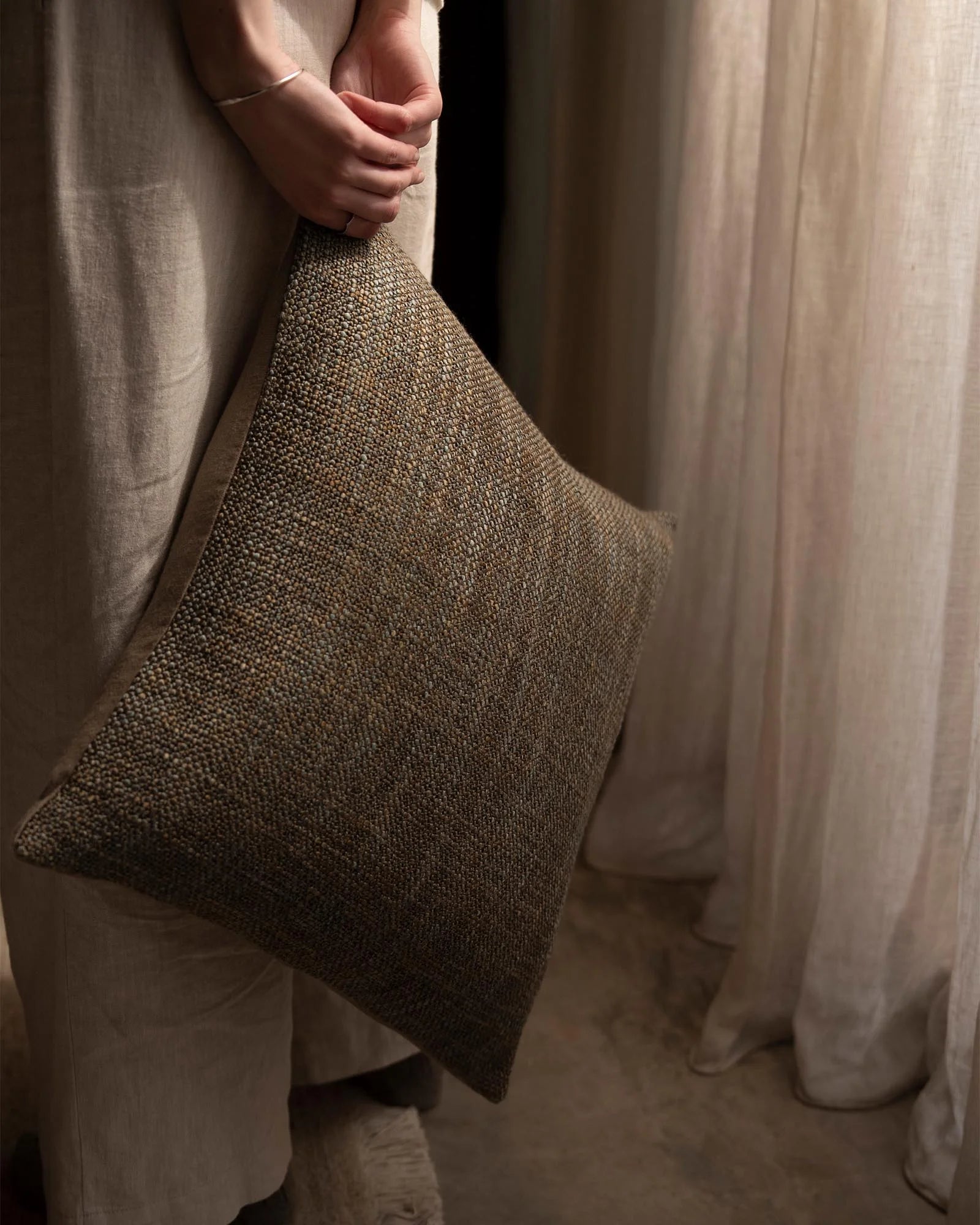 Hand holding textured olive-toned cushion against draped linen curtains in warm neutral palette, showcasing tactile woven weave and layered fabric styling
