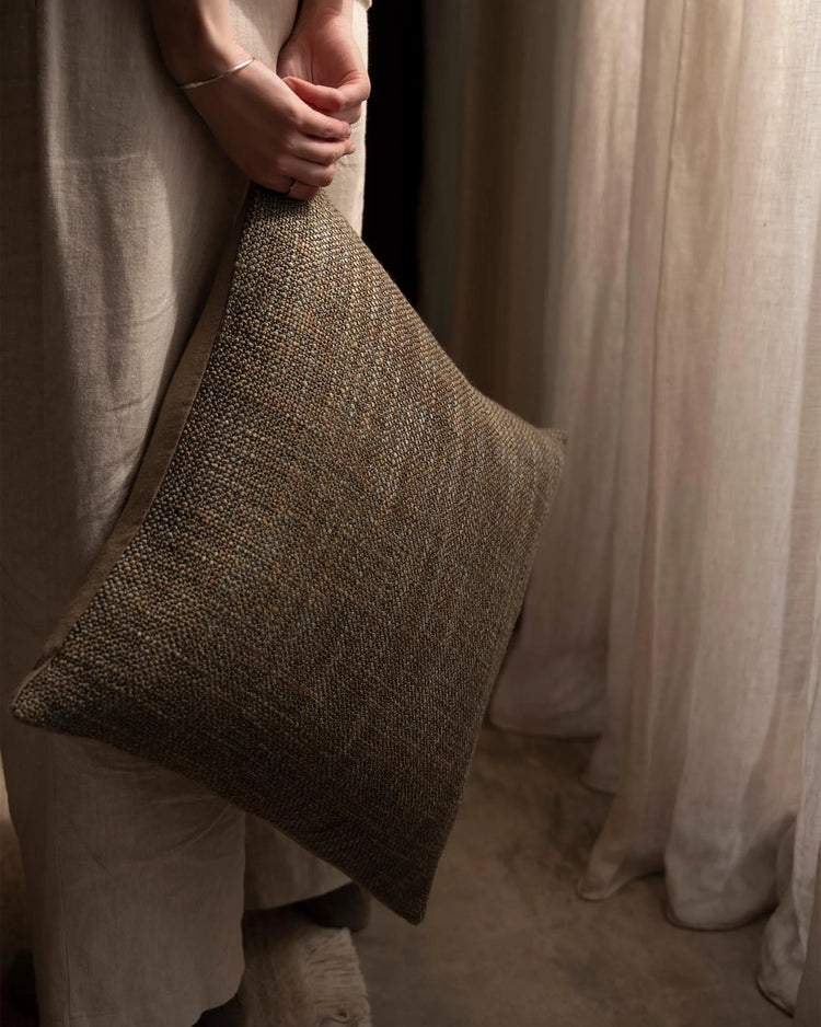 Hand holding textured olive-toned cushion against draped linen curtains in warm neutral palette, showcasing tactile woven weave and layered fabric styling