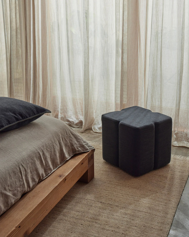 Minimalist bedroom with neutral linen curtains, oat-toned rug, and charcoal linen ottoman anchoring a wood-frame bed with taupe bedding