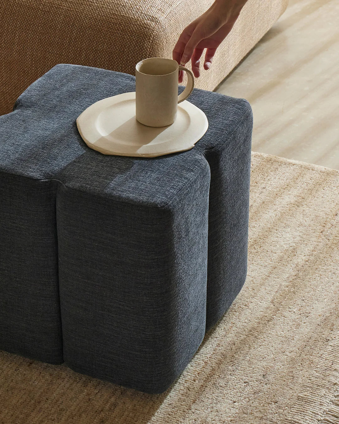 Slate-blue linen ottoman styled in warm neutral living room with cream ceramics, natural jute rug, and layered textural palette of woven furnishings