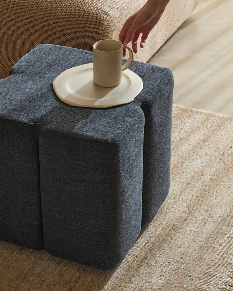 Slate-blue linen ottoman styled in warm neutral living room with cream ceramics, natural jute rug, and layered textural palette of woven furnishings
