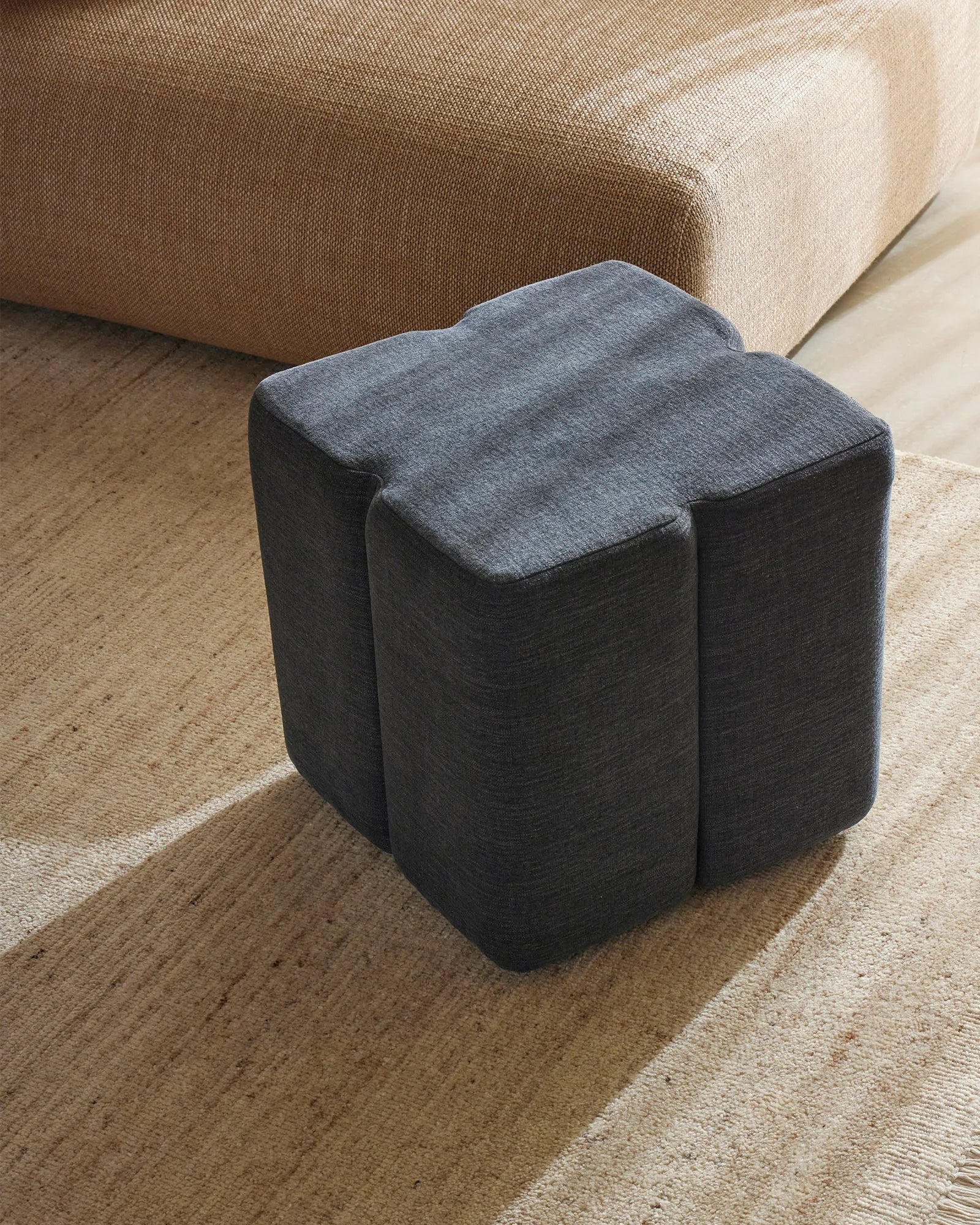 Charcoal linen ottoman beside beige wool rug, with warm neutral sofa backdrop, layered earth-tone interior styling