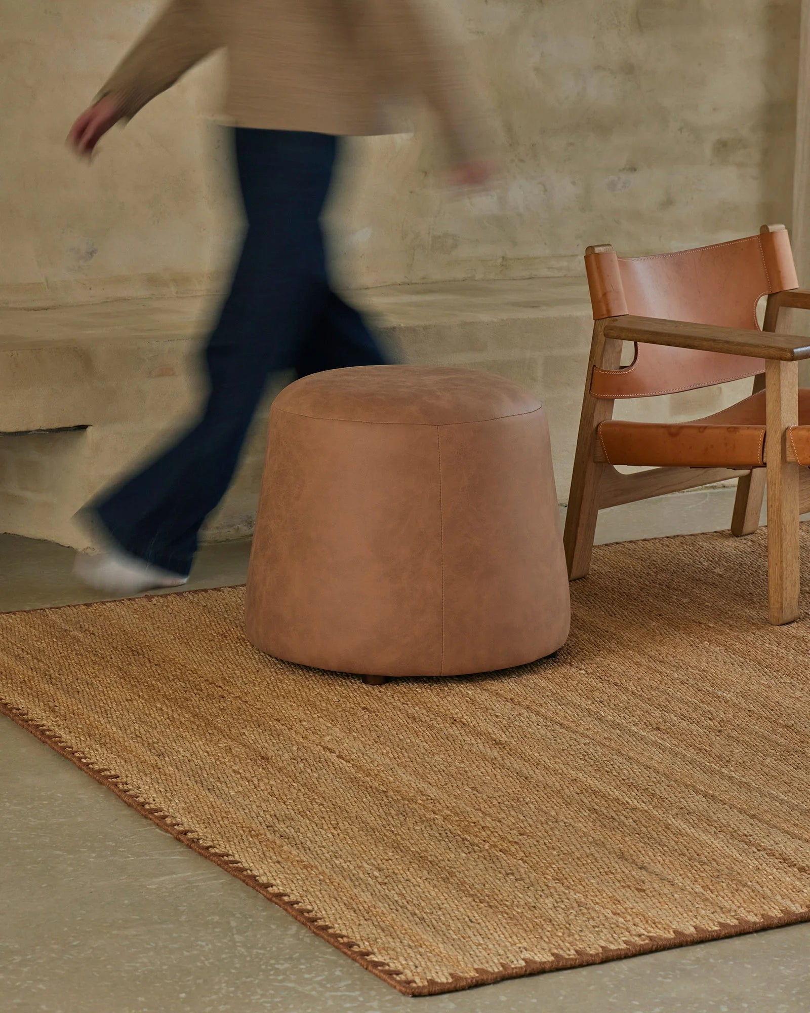 Warm neutral living space styled with Monroe Biscuit natural fibre rug, tan leather ottoman, and wooden chair against textured concrete wall backdrop