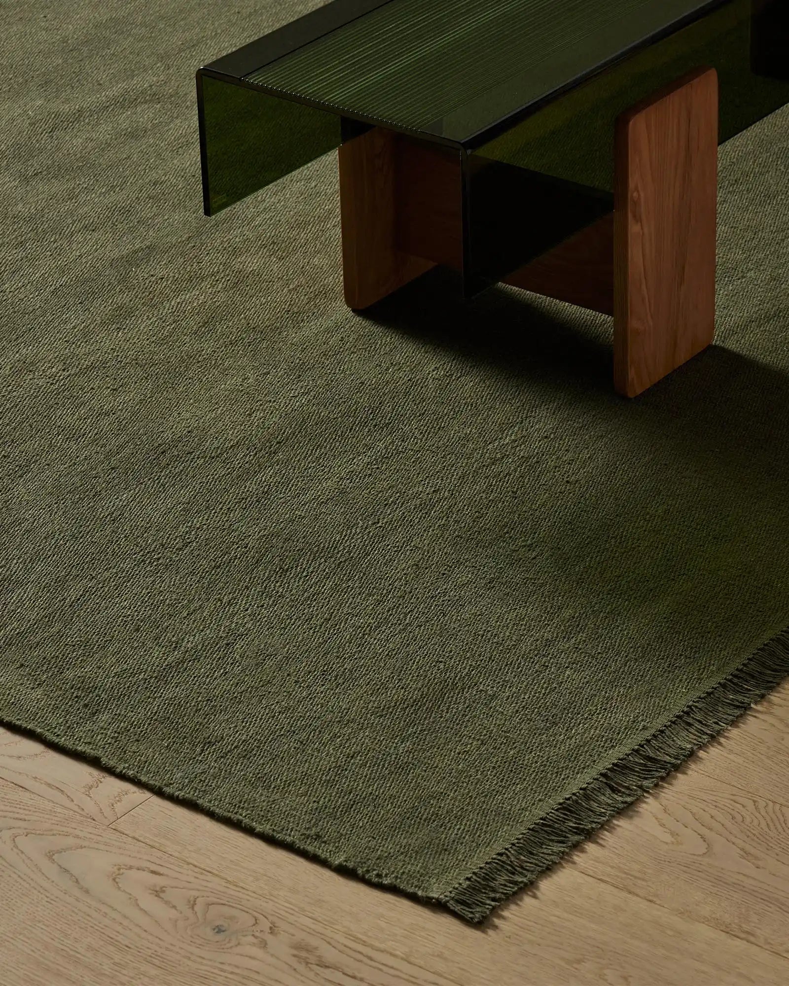 Sage green linen rug with subtle striped texture and fringed edge layered on light wooden flooring, styled under mid-century modern furniture in warm timber and forest tones
