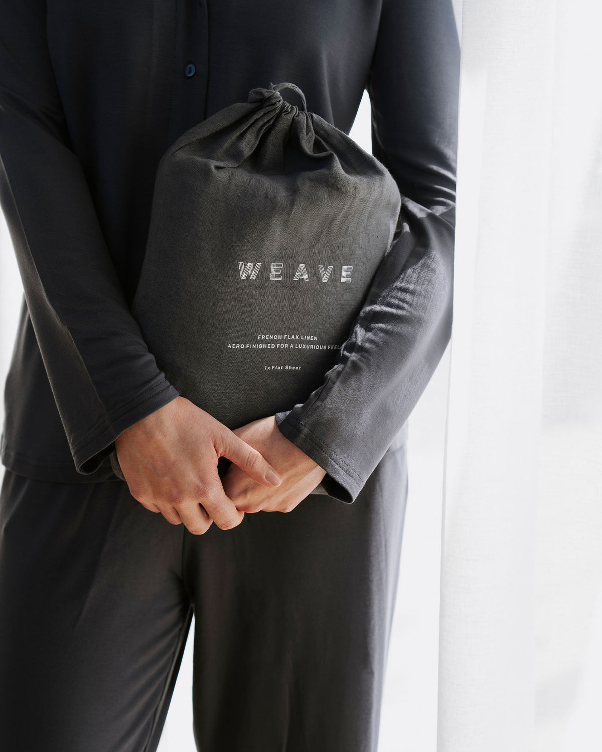 Charcoal linen bedding set in branded drawstring pouch, held against dark minimalist styling, showcasing soft European linen texture and monochromatic luxury aesthetic