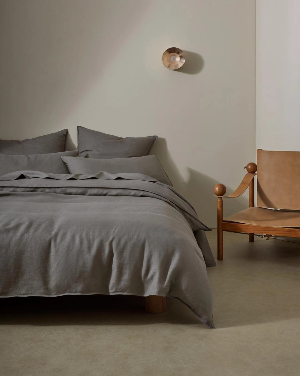 Minimalist bedroom with charcoal linen bedding layered on wooden bed frame, mid-century leather chair, neutral walls, warm natural light, Scandinavian-inspired interior styling