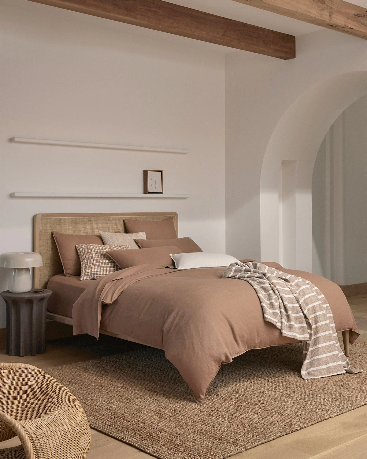 Warm minimalist bedroom with biscuit linen bedding, rattan headboard, and terracotta throw layered over neutral bedding, styled with geometric cushion and natural textures