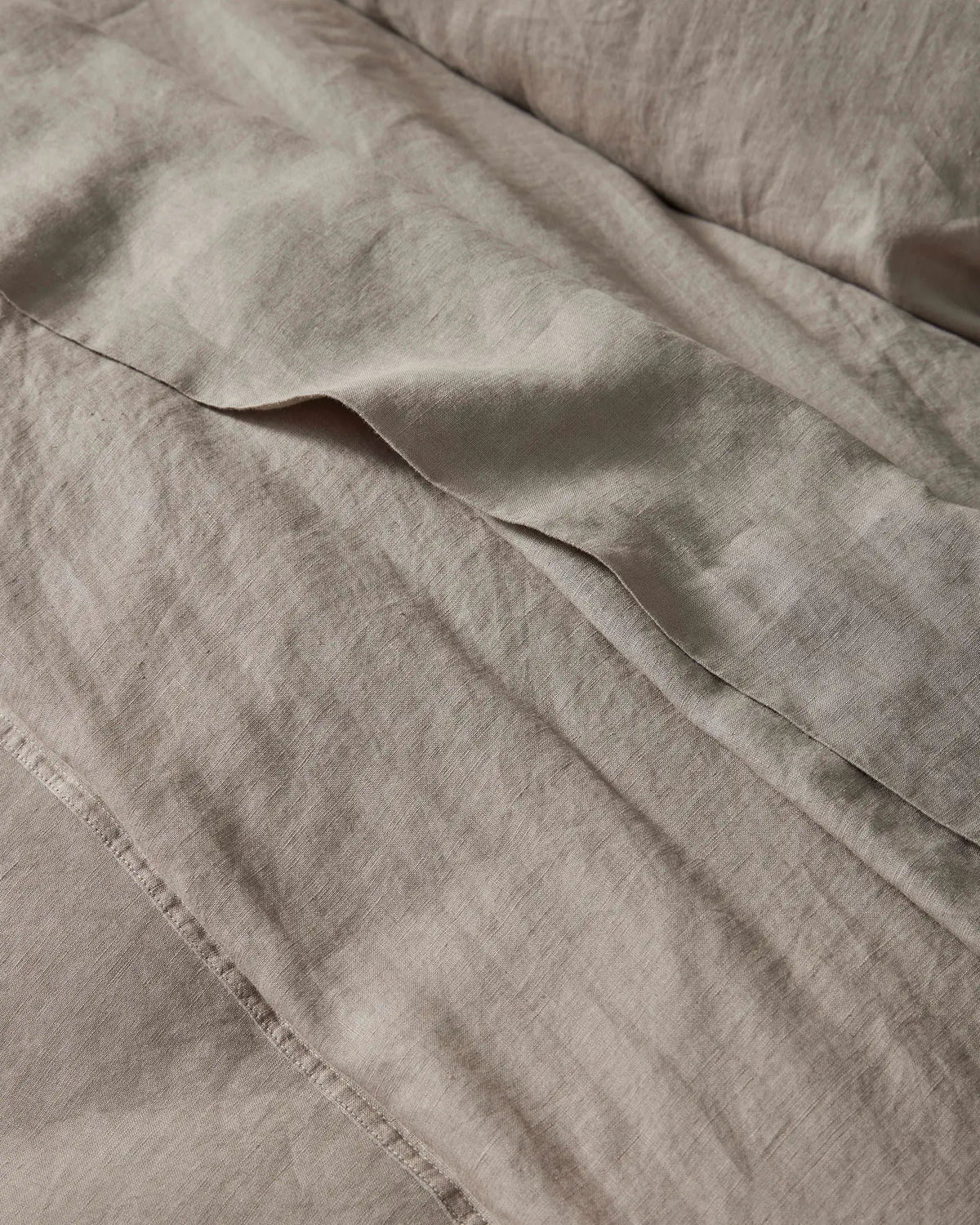 Neutral linen bedding in warm pebble tones with soft texture and gentle folds, showcasing the Ravello Flat Sheet's lightweight European linen weave and relaxed, undone aesthetic