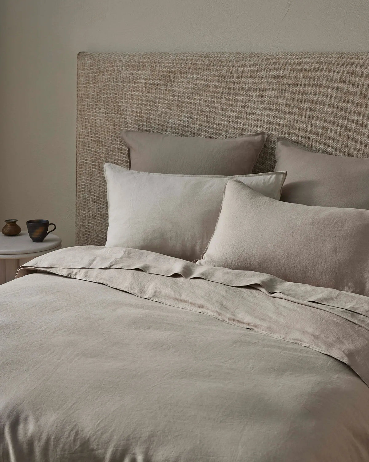 Neutral minimalist bedroom styled with layered Ravello flat sheet in soft pebble tones, textured linen bedding, and upholstered headboard, warm ambient lighting