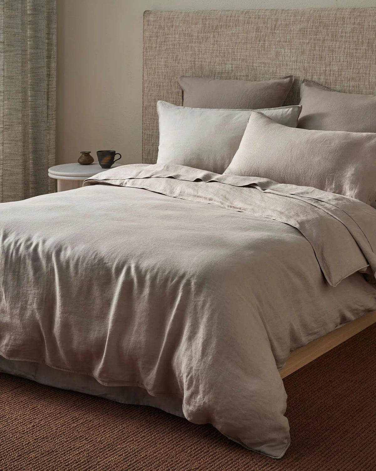 Neutral bedroom with layered linen bedding in warm sand and pebble tones, textured headboard, and earthy minimalist styling with ceramic accessories on wooden nightstand.