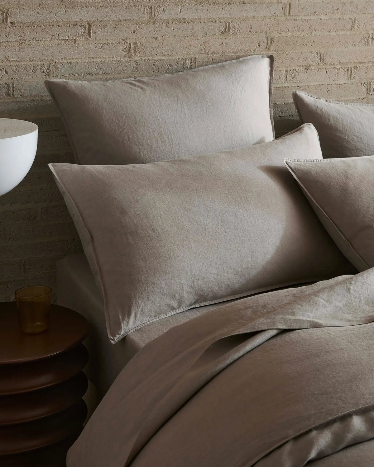 Neutral linen bedding styled on a bed with textured brick wall, featuring layered Ravello flat sheet in warm pebble tones and soft furnishings for minimalist bedroom aesthetic