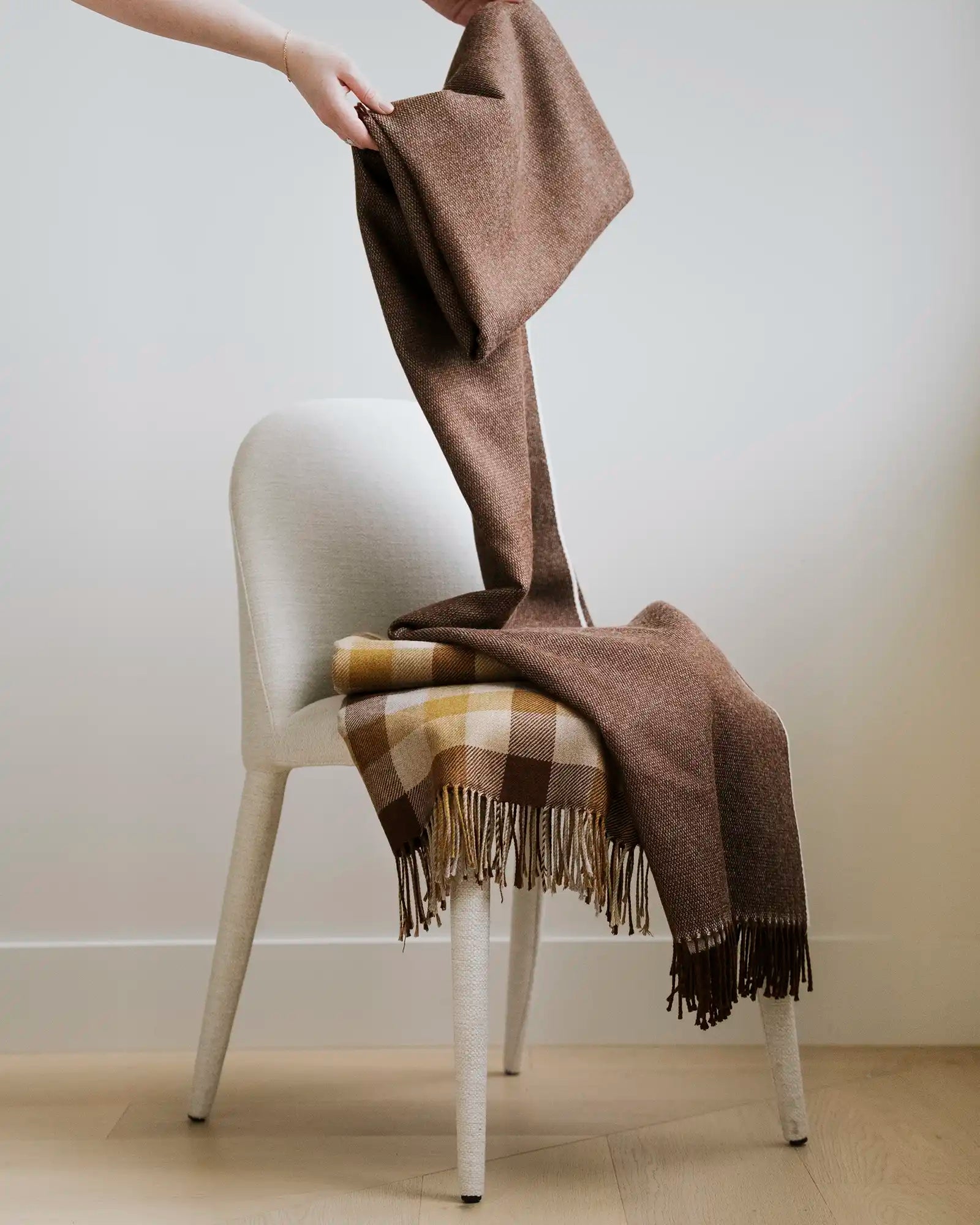 Neutral modern chair layered with warm brown wool throws and a gold check pattern throw with fringe trim, creating an earthy autumn palette against pale wooden flooring