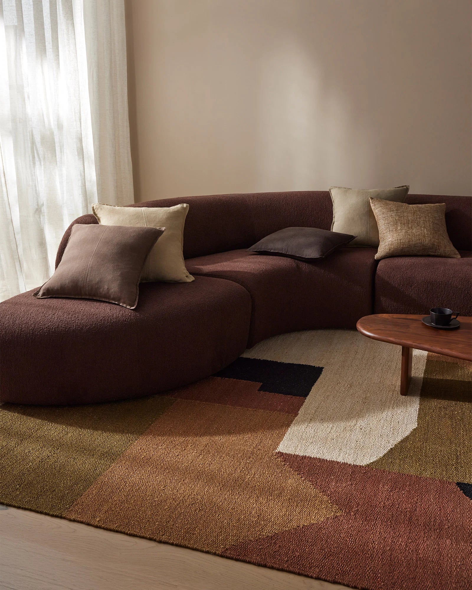 Warm minimalist living room with chocolate sectional, cream and tan scatter cushions, geometric terracotta and cream jute rug, timber coffee table, and soft linen curtains