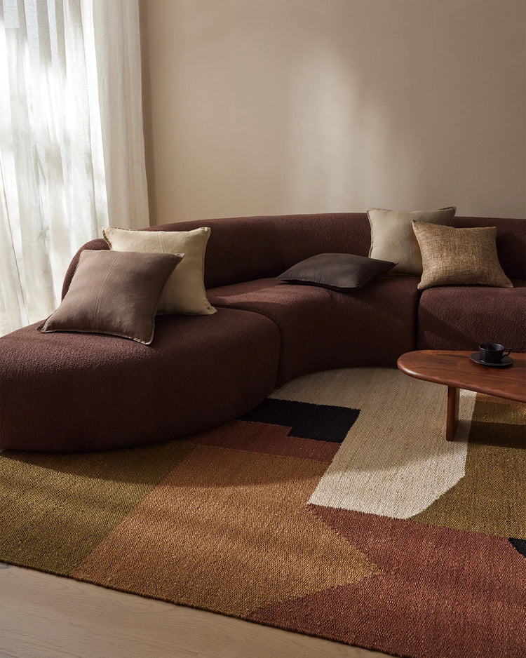 Warm minimalist living room with chocolate sectional, cream and tan scatter cushions, geometric terracotta and cream jute rug, timber coffee table, and soft linen curtains