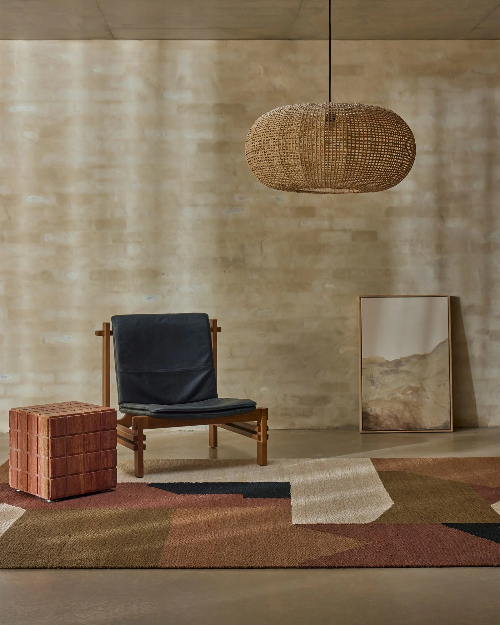 Earthy minimalist interior with geometric terracotta and neutral rug, timber lounge chair, woven pendant and brick-textured wall backdrop