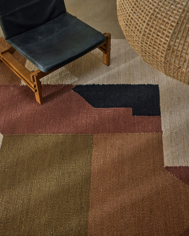 Earthy-toned rug in terracotta, olive and charcoal with geometric colour-block pattern, styled in modern living room with mid-century wooden chair and woven pouf accent