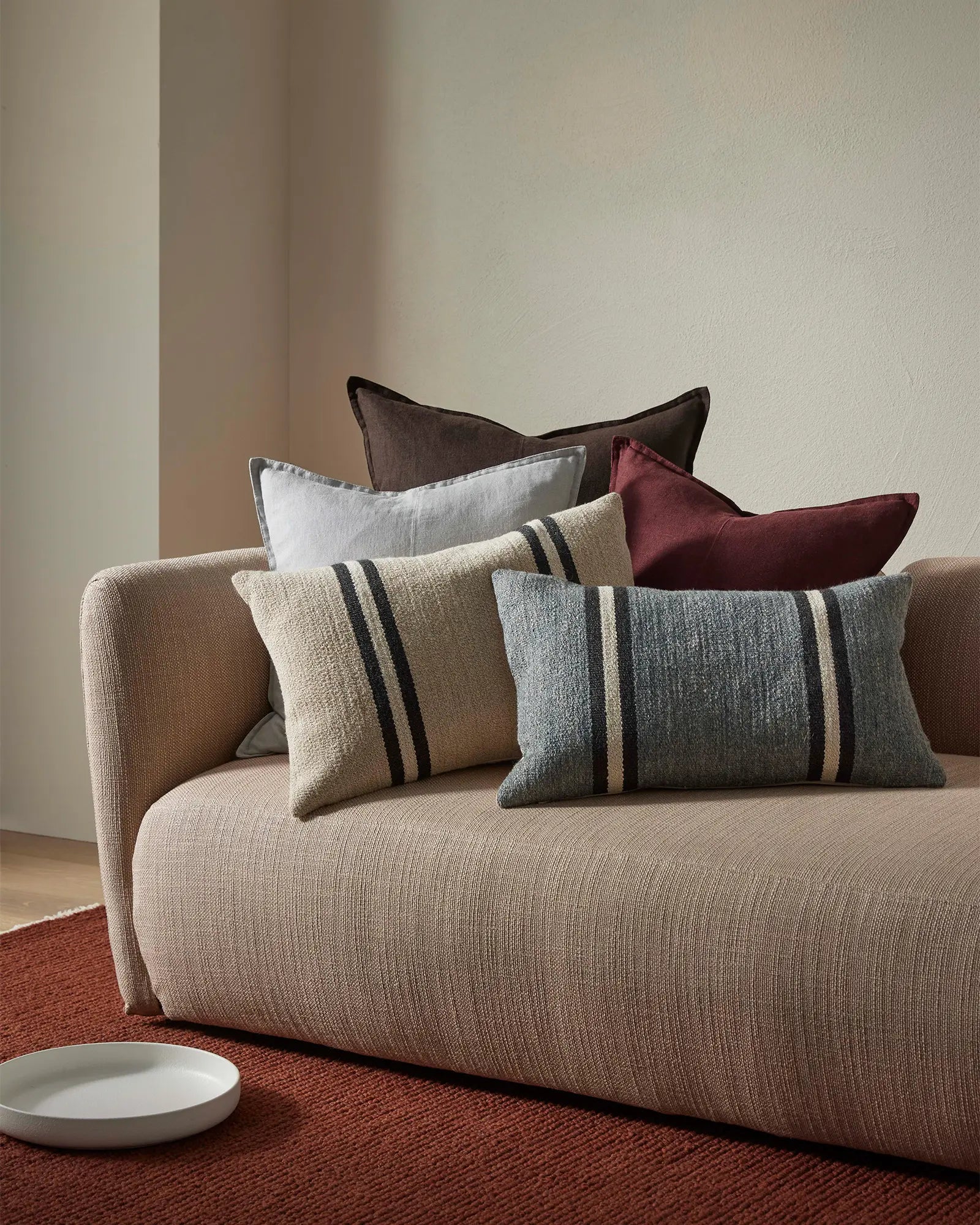 Neutral linen sofa layered with striped wool cushions in grey and cream, burgundy and charcoal accents, warm minimalist interior styling