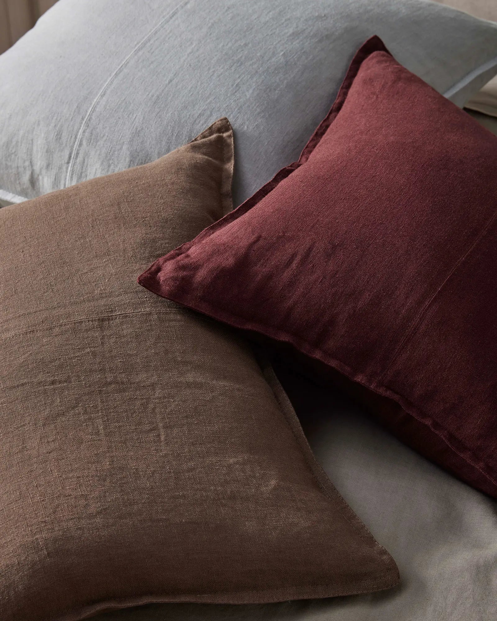 Layered linen cushions in warm mocha and deep garnet tones on a pale grey upholstered chair, creating an earthy, textural palette with natural linen weave detail