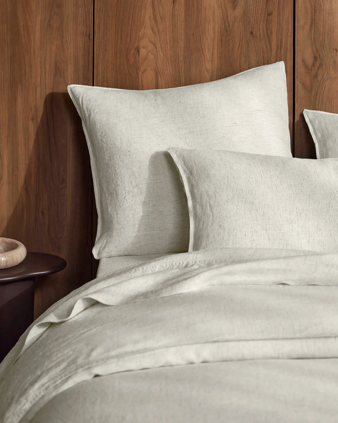 Neutral linen bedding styled on a bed against warm timber paneling, featuring cream Sova pillows and sheets with subtle pinstripe texture