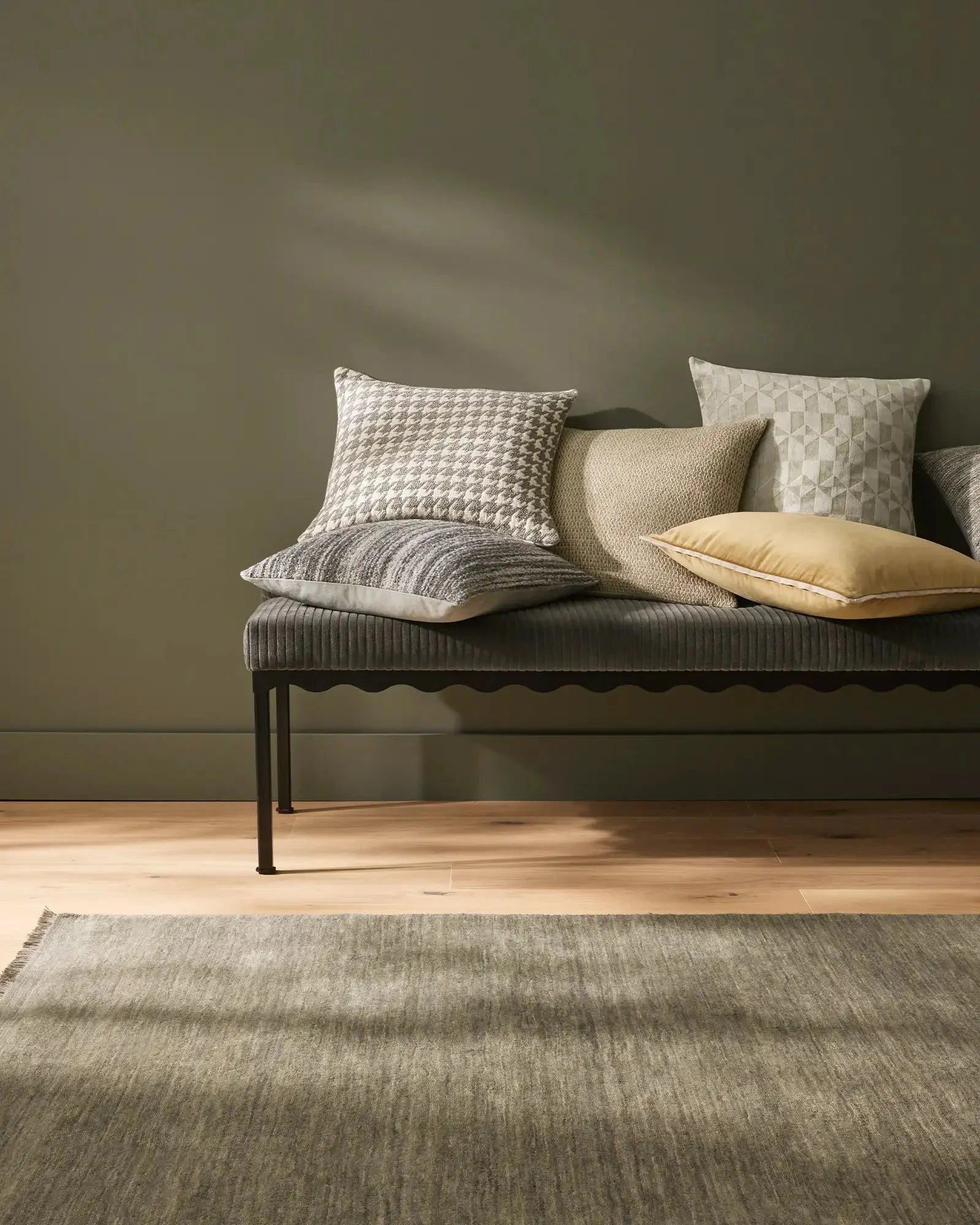 Sage green bedroom styled with textured throw cushions in cream, grey and pale yellow on a ribbed bench, warm timber flooring and neutral area rug creating calm, minimalist aesthetic
