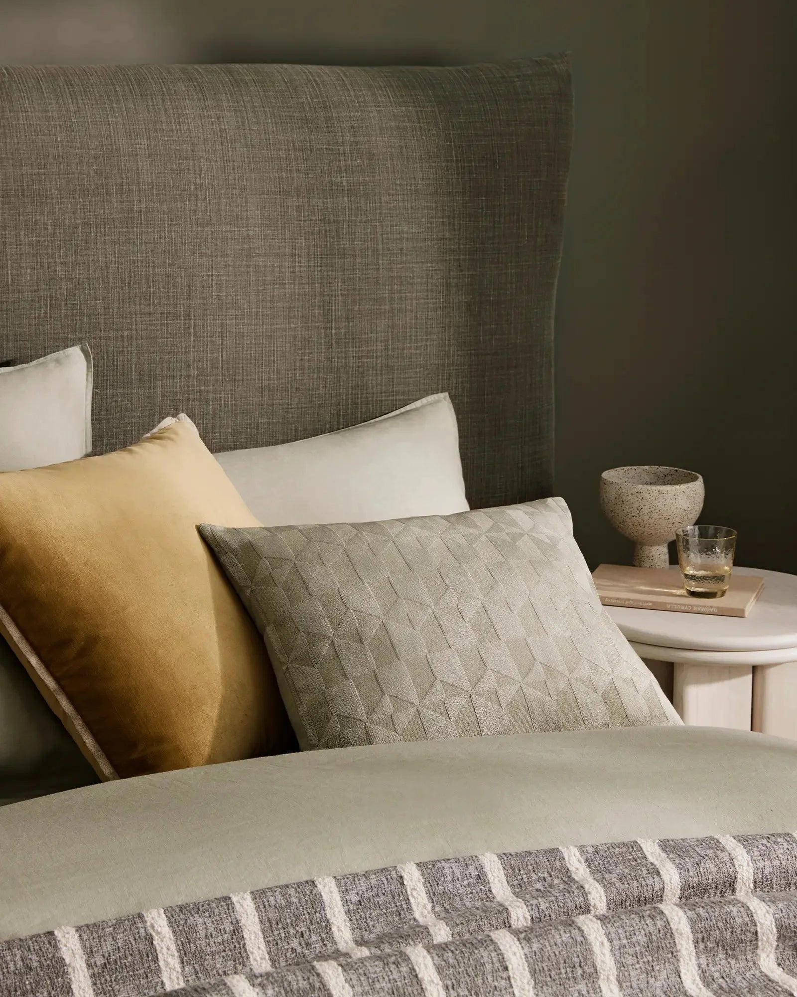 Neutral bed styled with sage linen flat sheet, golden cushion, quilted sage cushion and striped throw, grey upholstered headboard, minimalist bedside styling
