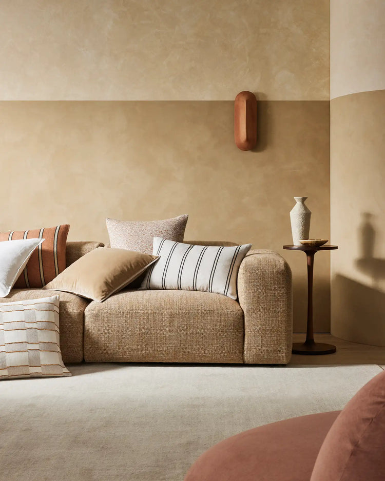 Warm neutral living room with beige sofa and striped scatter cushions in terracotta and cream tones, layered textures including linen and woven fabrics, minimalist aesthetic