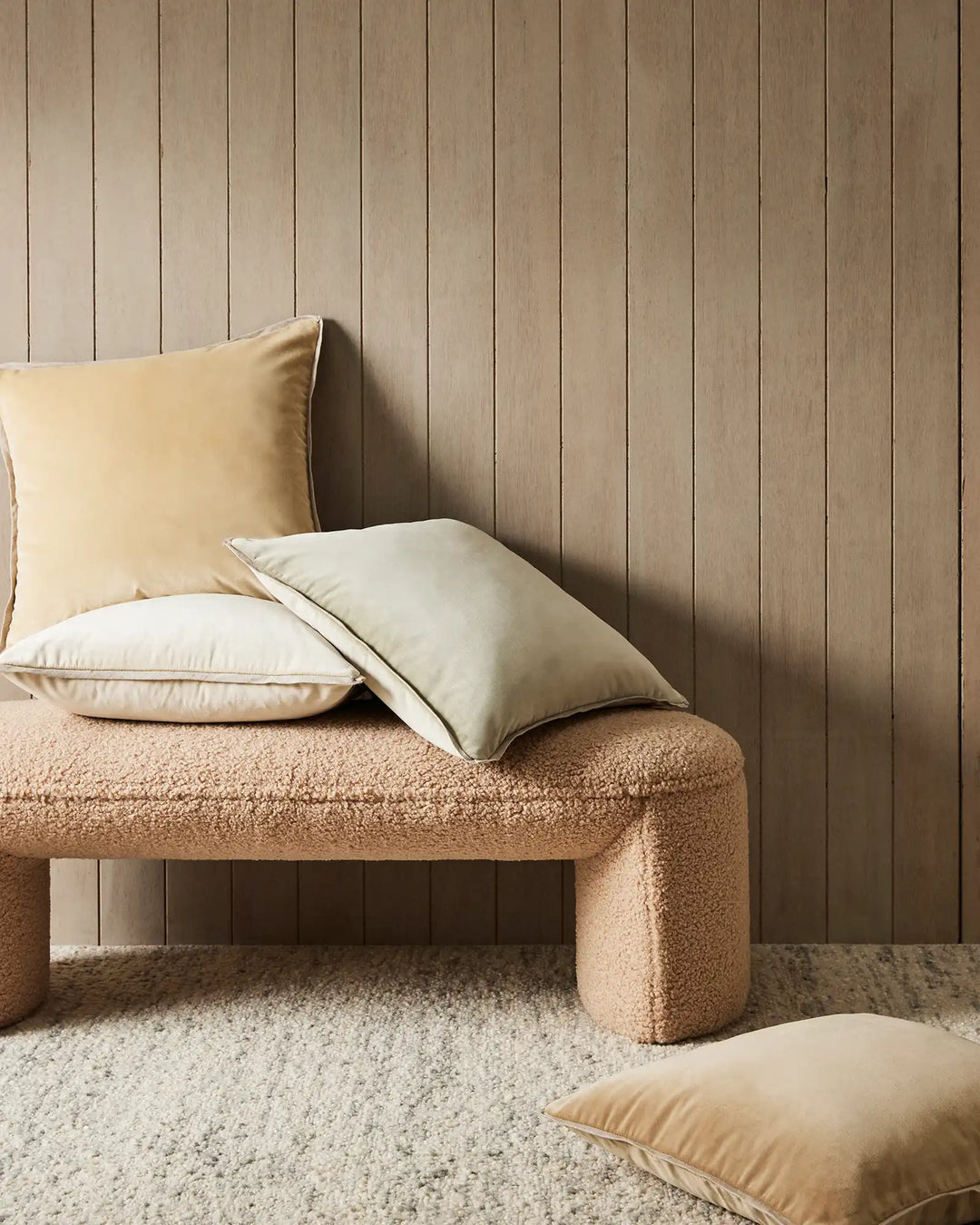 Neutral minimalist bedroom scene with cream and sage cushions styled on textured boucle bench, vertical wood paneling backdrop, layered earth tones and natural fibres