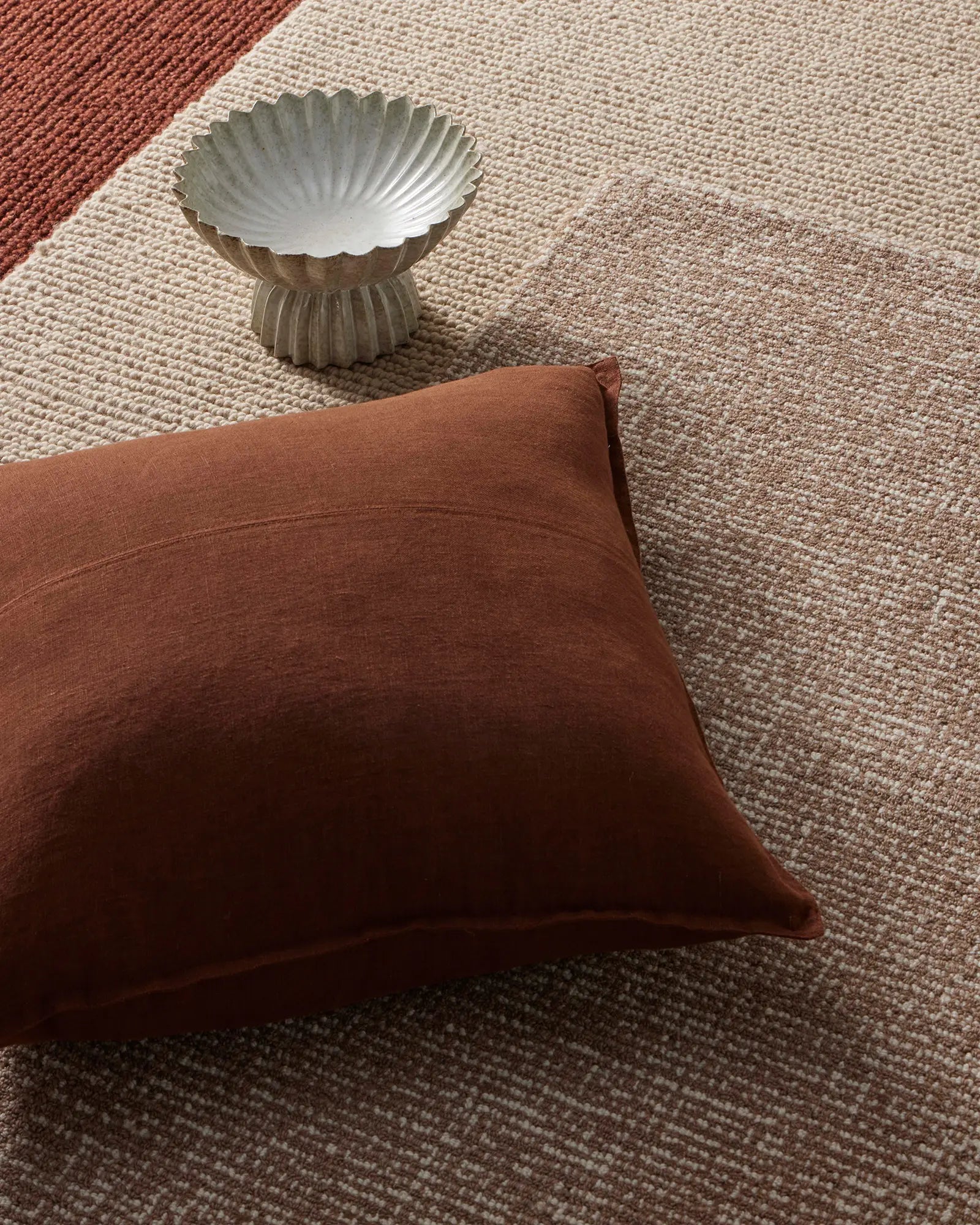 Rust and cream linen cushion and woven rugs layered on natural textiles, with sculptural ceramic bowl, earthy tonal palette styling