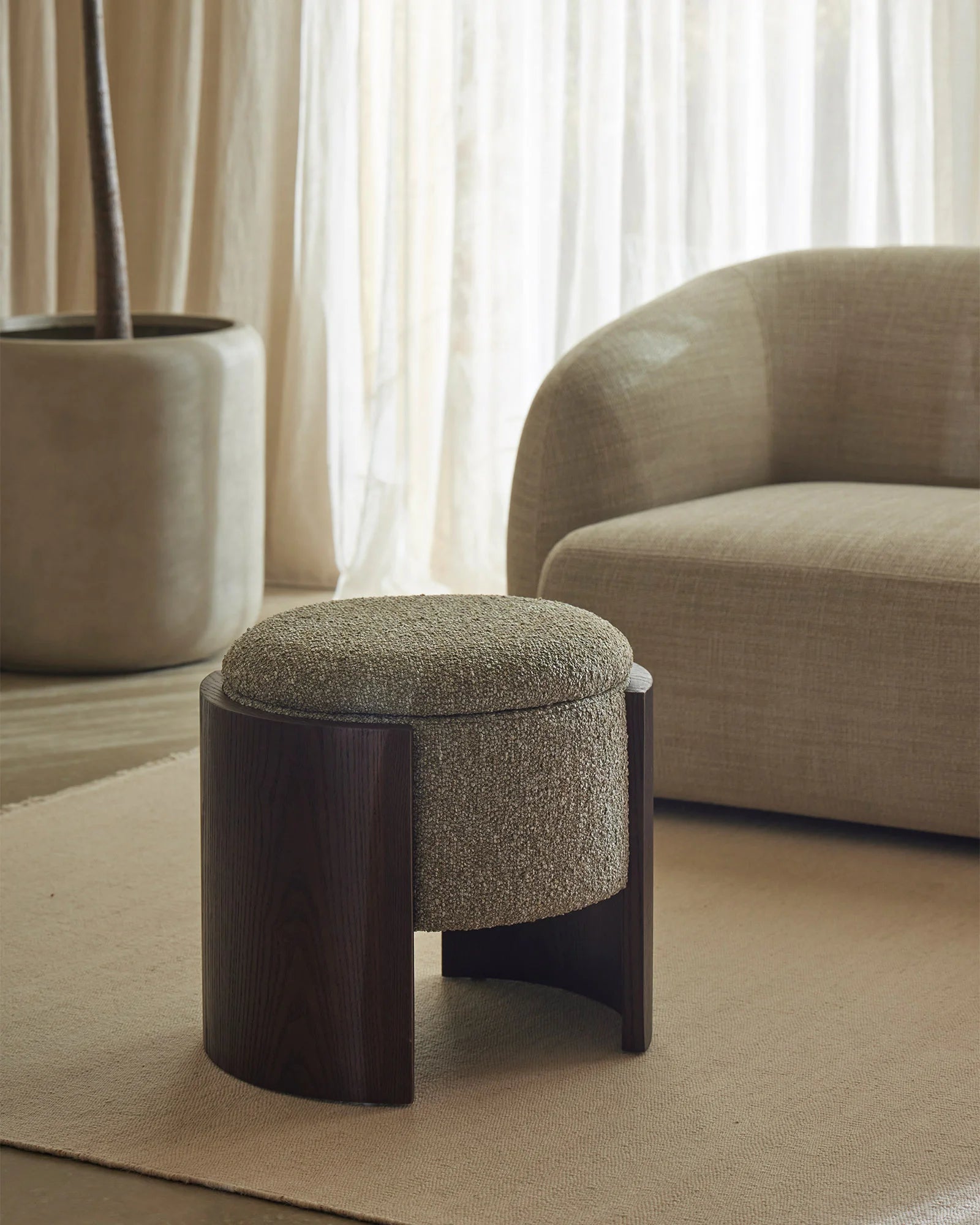 Minimalist living room with neutral palette, featuring warm beige linen armchair, moss green textured ottoman with dark wood base, and cream wool area rug, styled with sheer curtains and potted plant.