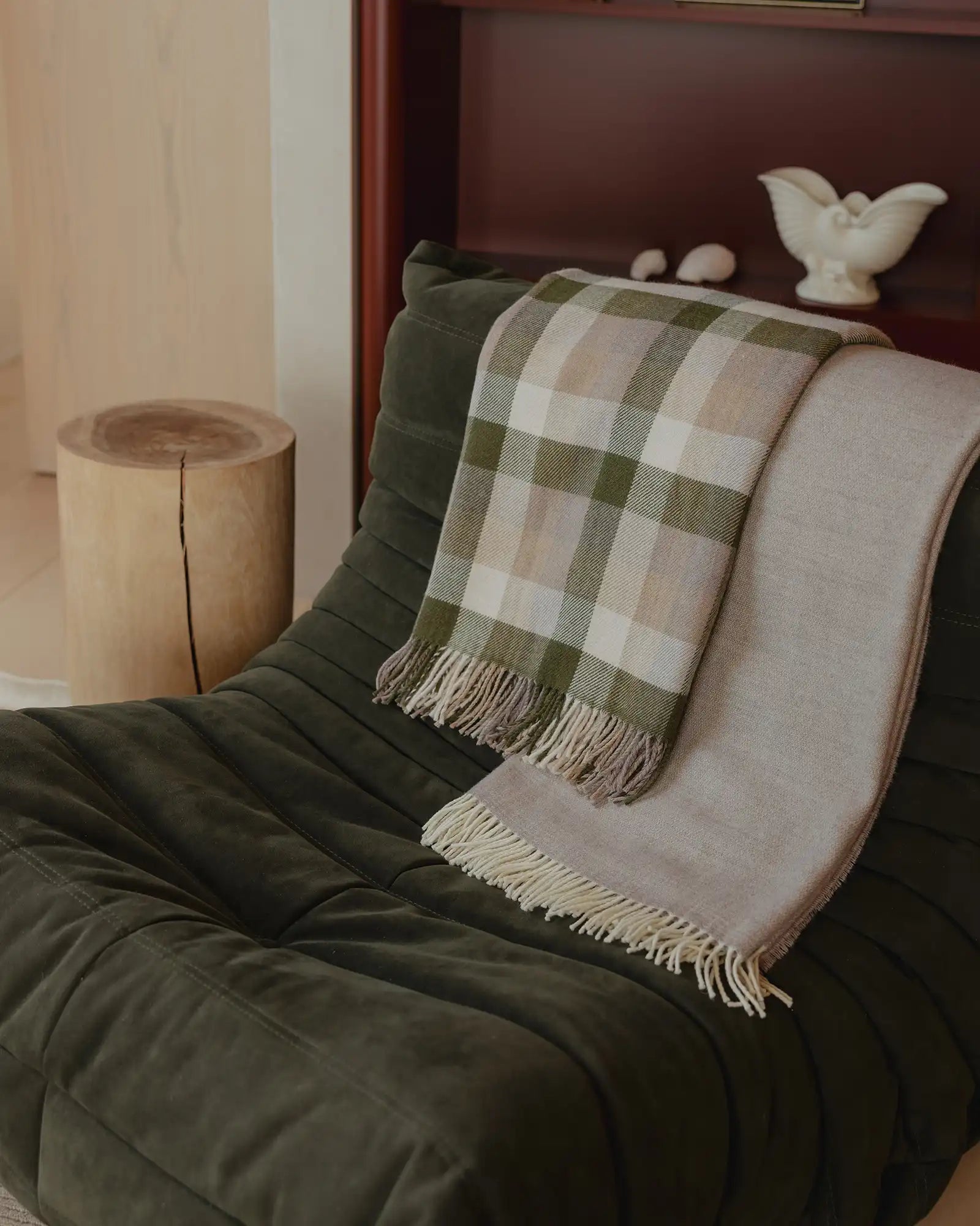 Sage and cream checked wool throw layered with natural linen throw on dark olive quilted bedding, minimalist wooden stool beside bed, earthy autumn palette