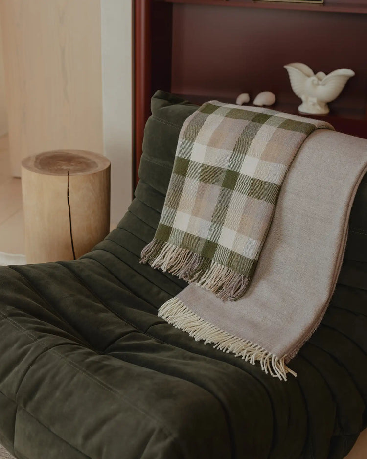 Sage and cream checked wool throw layered with natural linen throw on dark olive quilted bedding, minimalist wooden stool beside bed, earthy autumn palette