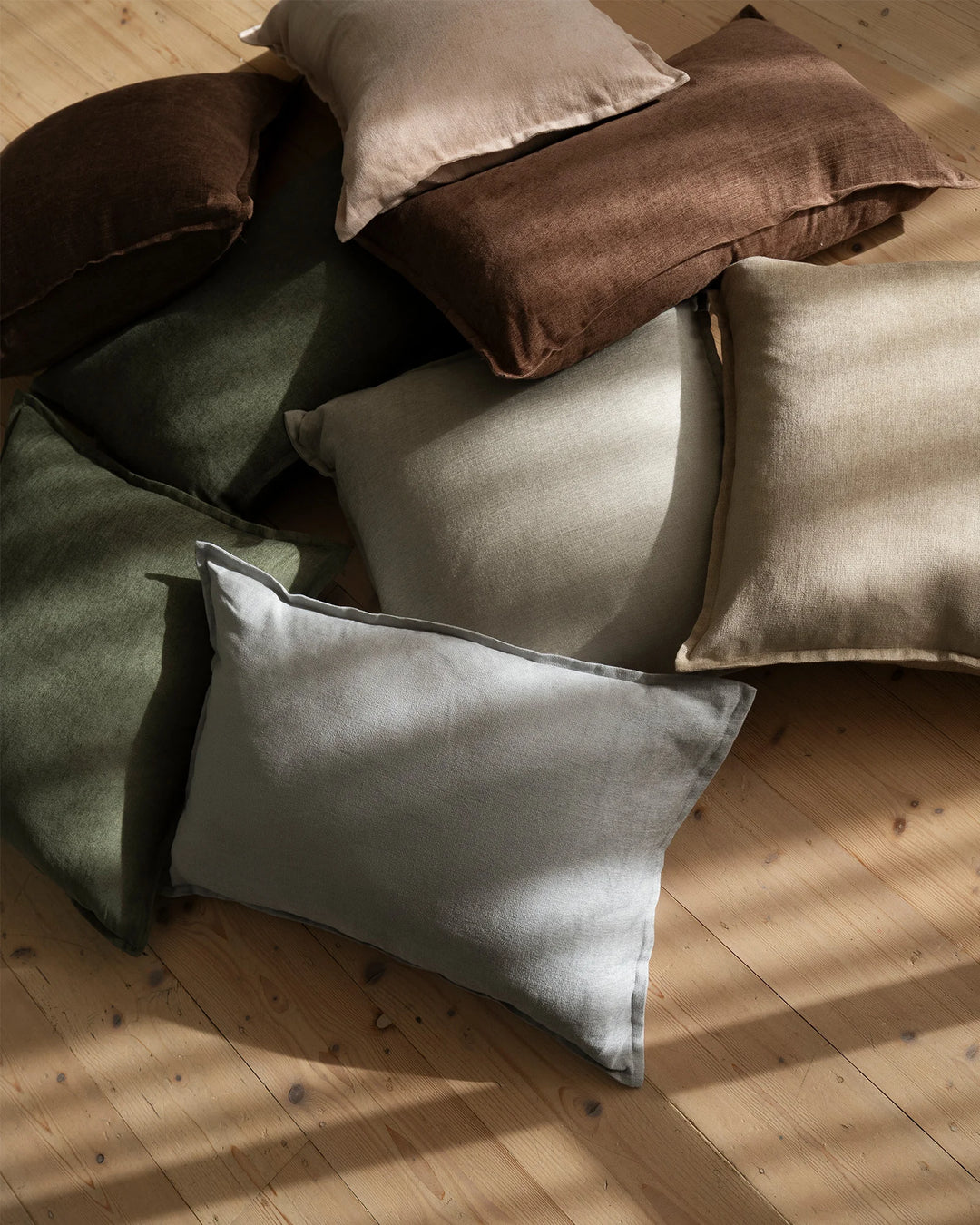 Overhead view of linen viscose blend cushions in earthy tones—sage, olive, toffee, ecru, and breeze—arranged on warm timber flooring, showcasing natural texture and neutral palette styling