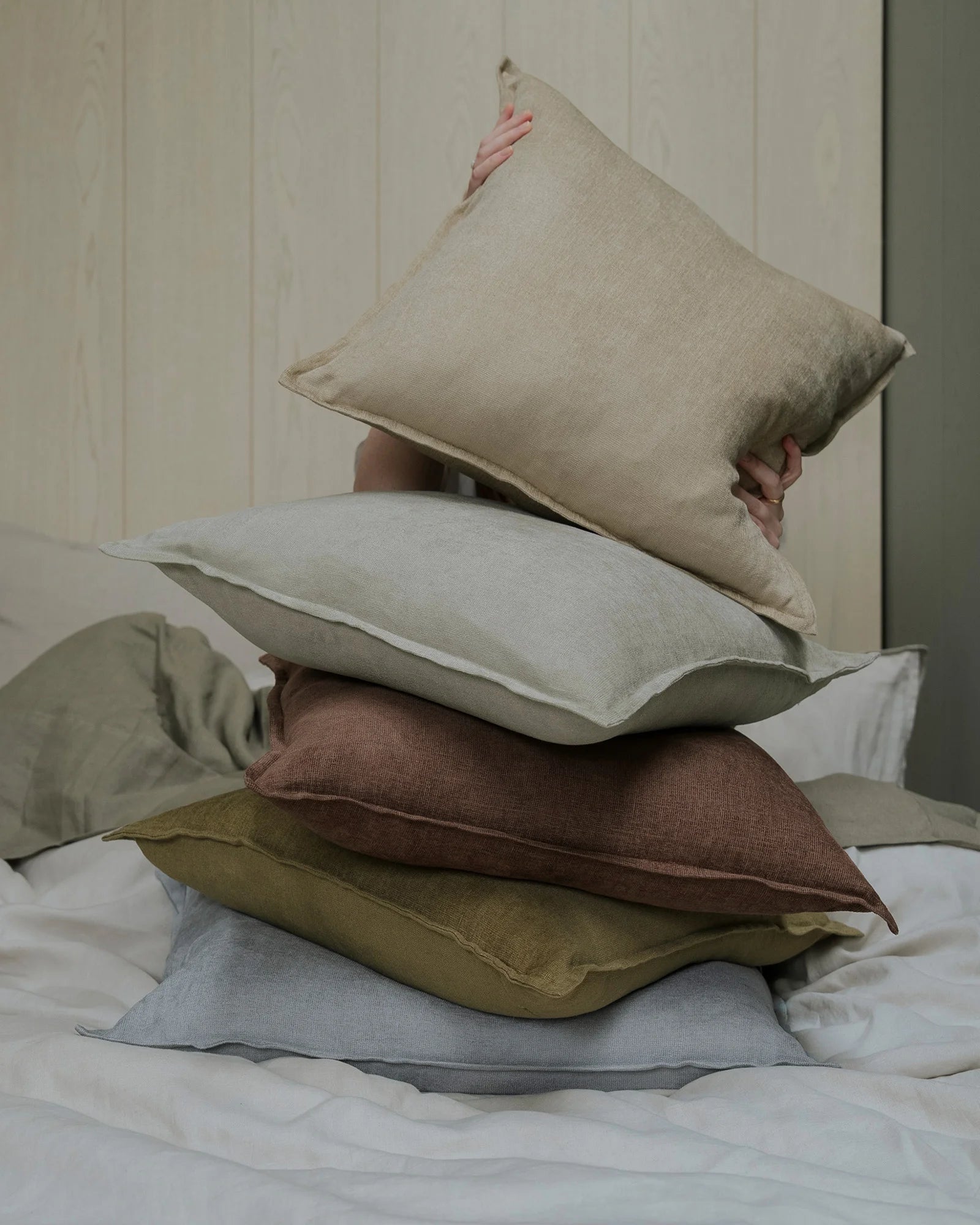 Stacked Fiore cushions in neutral earth tones—sage, toffee, ecru, and breeze—showcase linen viscose blend textures against white panelled walls and bedding