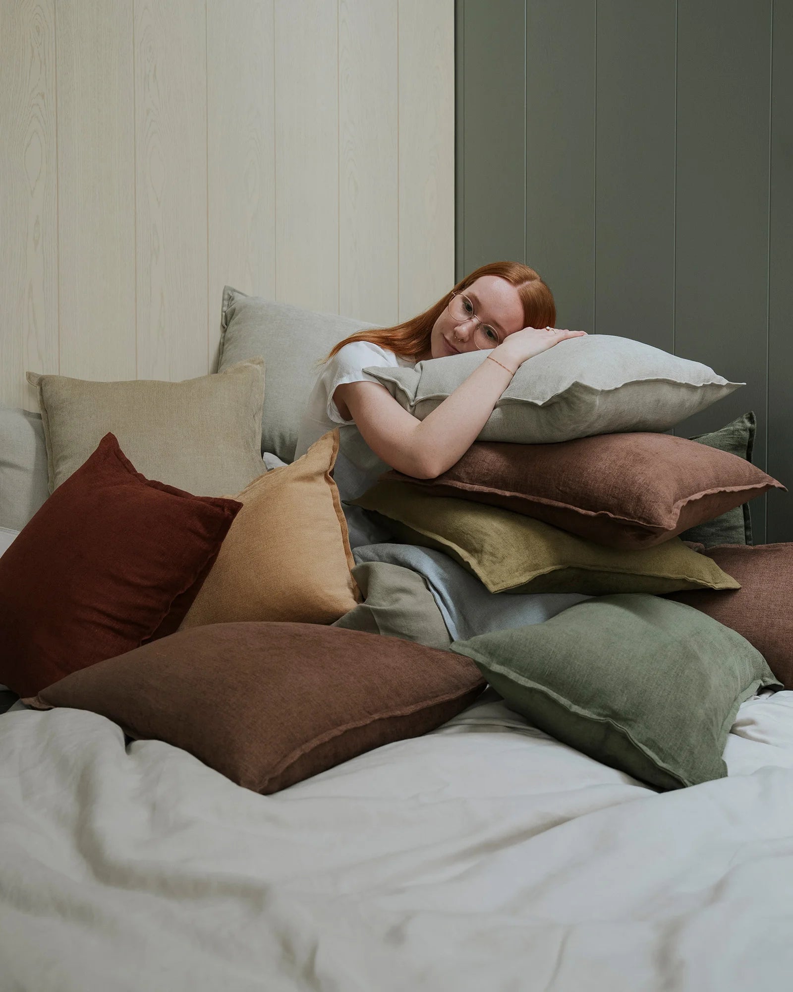 Cosy bedroom scene with stacked Fiore cushions in earthy tones—sage, olive, honey, rosewood and ecru—against panelled green and white walls, natural linen textures layered for comfort and warmth