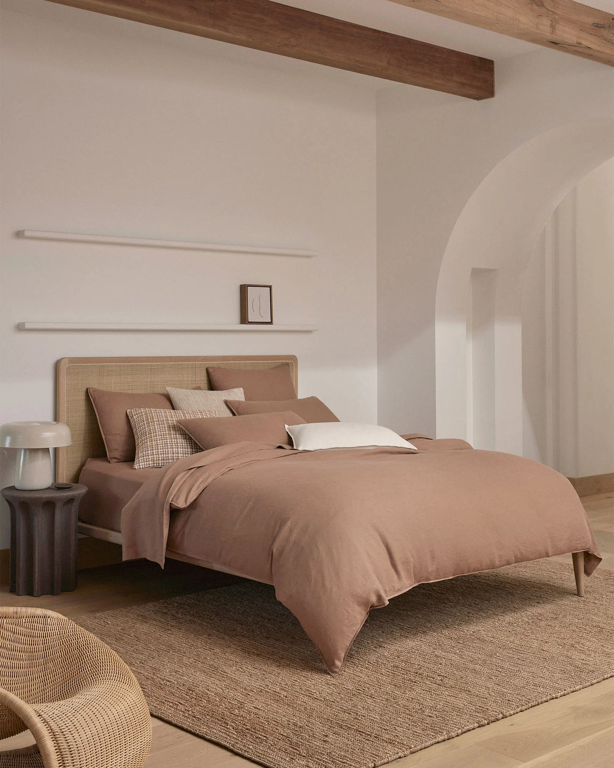 Minimalist bedroom with warm biscuit bedding, rattan headboard, and natural linen textures layered with neutral tones, timber beams, and woven rugs in a serene coastal aesthetic