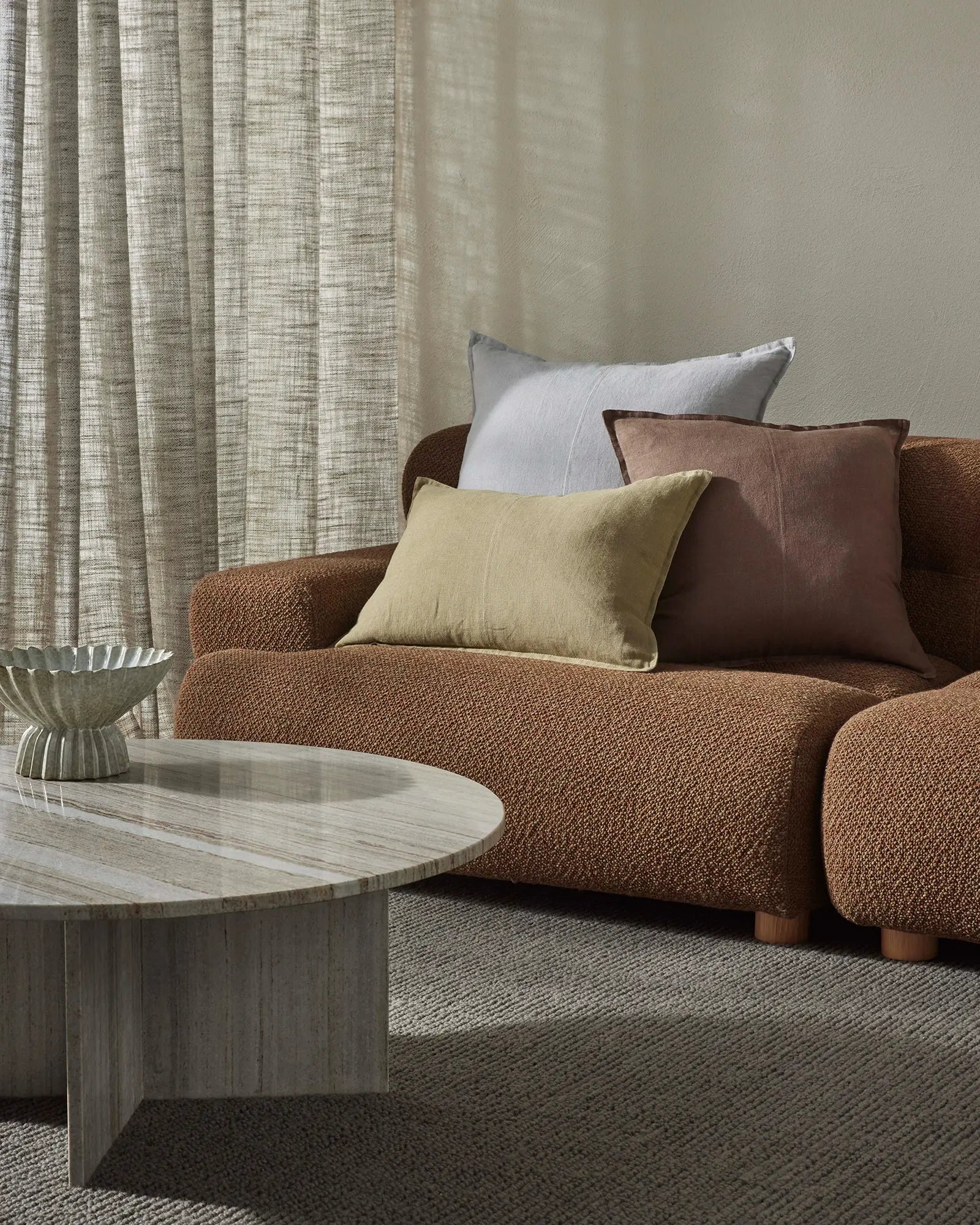 Warm neutral living room with terracotta sofa, stacked cushions in cream and chocolate tones, textured linen curtains, pale wood side table, and Fabien Echo wool rug in soft grey-beige