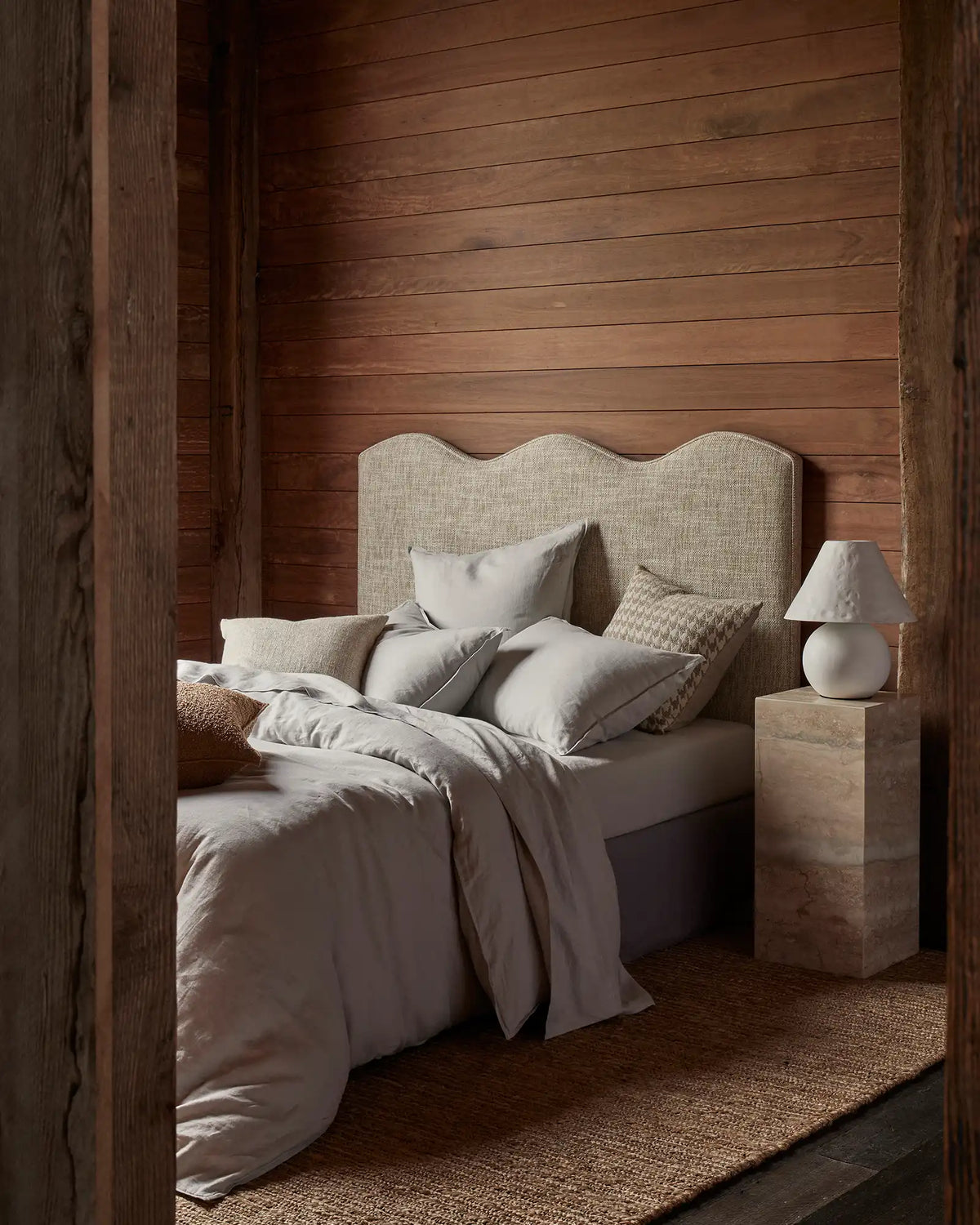 Warm timber bedroom with neutral layered bedding, cream upholstered headboard, and soft grey-toned linen sheets creating a calm, minimalist retreat with natural textures and earthy palette
