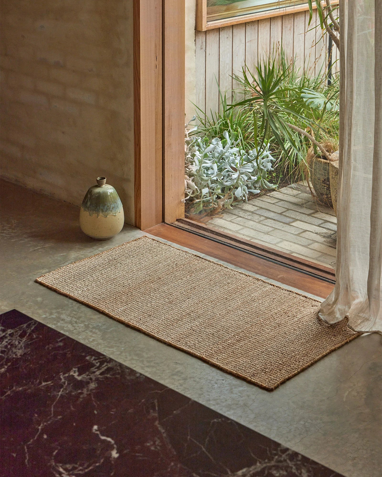 Neutral entryway styled with natural jute door mat, cream linen curtain, and ceramic vessel, opening to coastal garden with green foliage and whitewashed timber decking